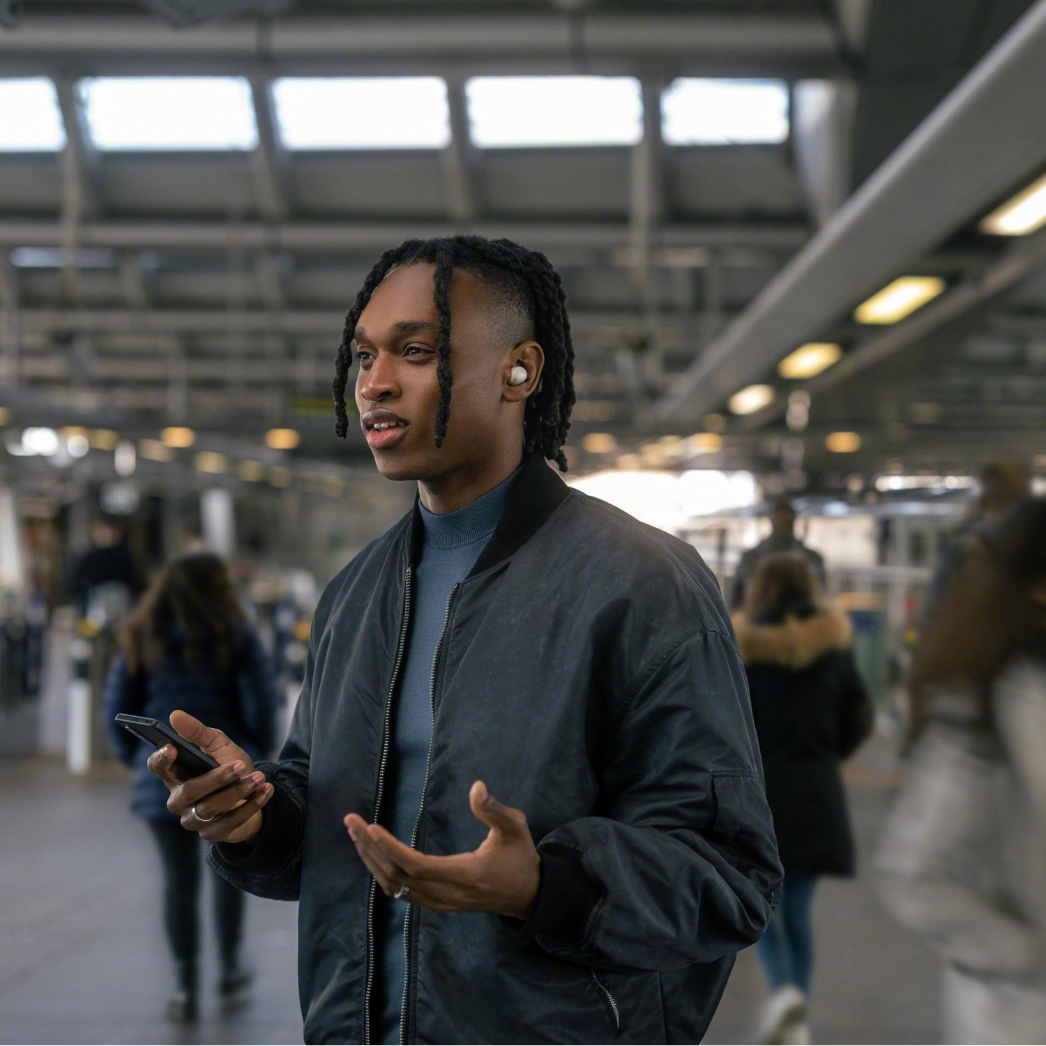Een man met dreadlocks houdt een telefoon vast en gebaart in een stationsachtige omgeving. Mensen zijn wazig op de achtergrond.