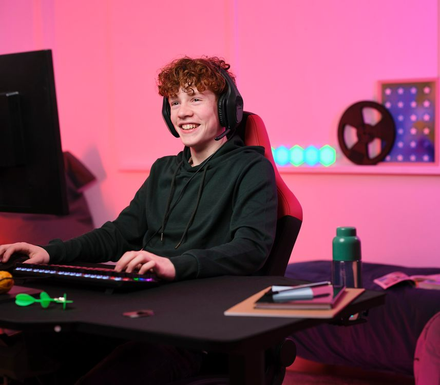 Egy fiatal gamer fejhallgatóval mosolyog, miközben számítógépen játszik. A szoba rózsaszínben világít.