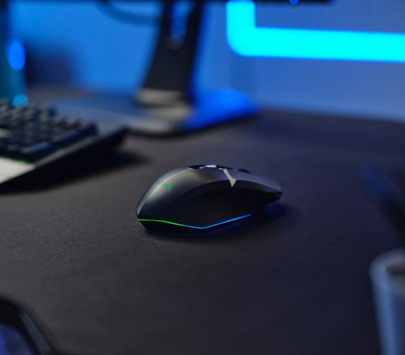 Fekete gamer egér fekete asztalon. Billentyűzet és monitor a homályos háttérben.