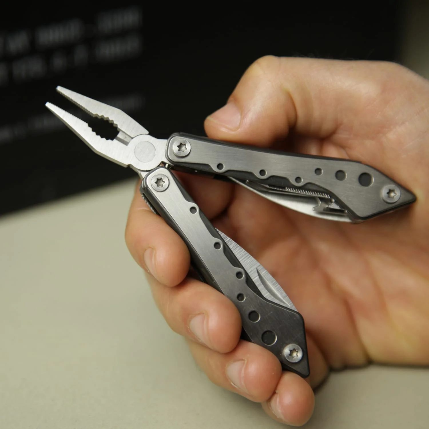 Eine Hand hält ein silbernes Multitool mit Zange und Messer, teilweise geöffnet, auf einer Oberfläche.
