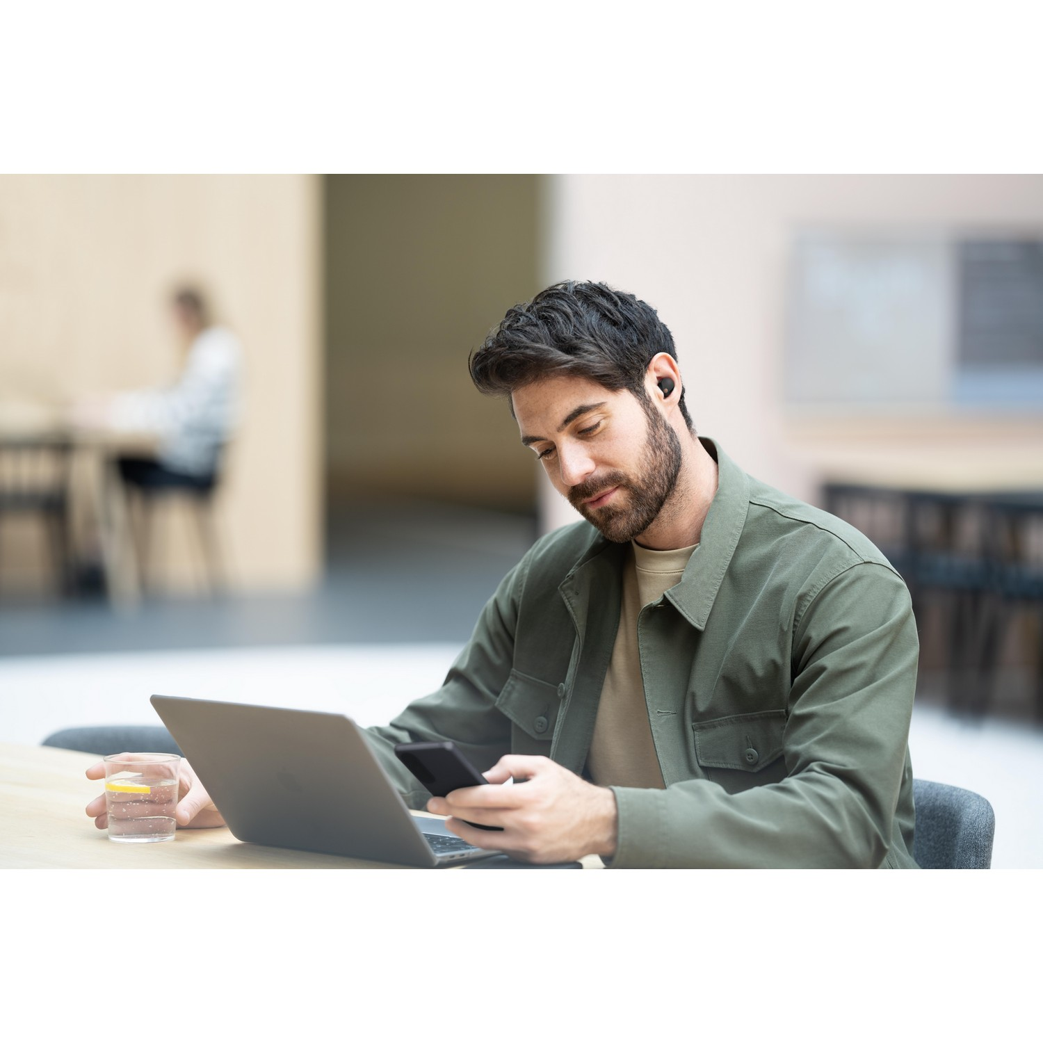 Een man die een laptop gebruikt, een telefoon vasthoudt en een glas water op een bureau.