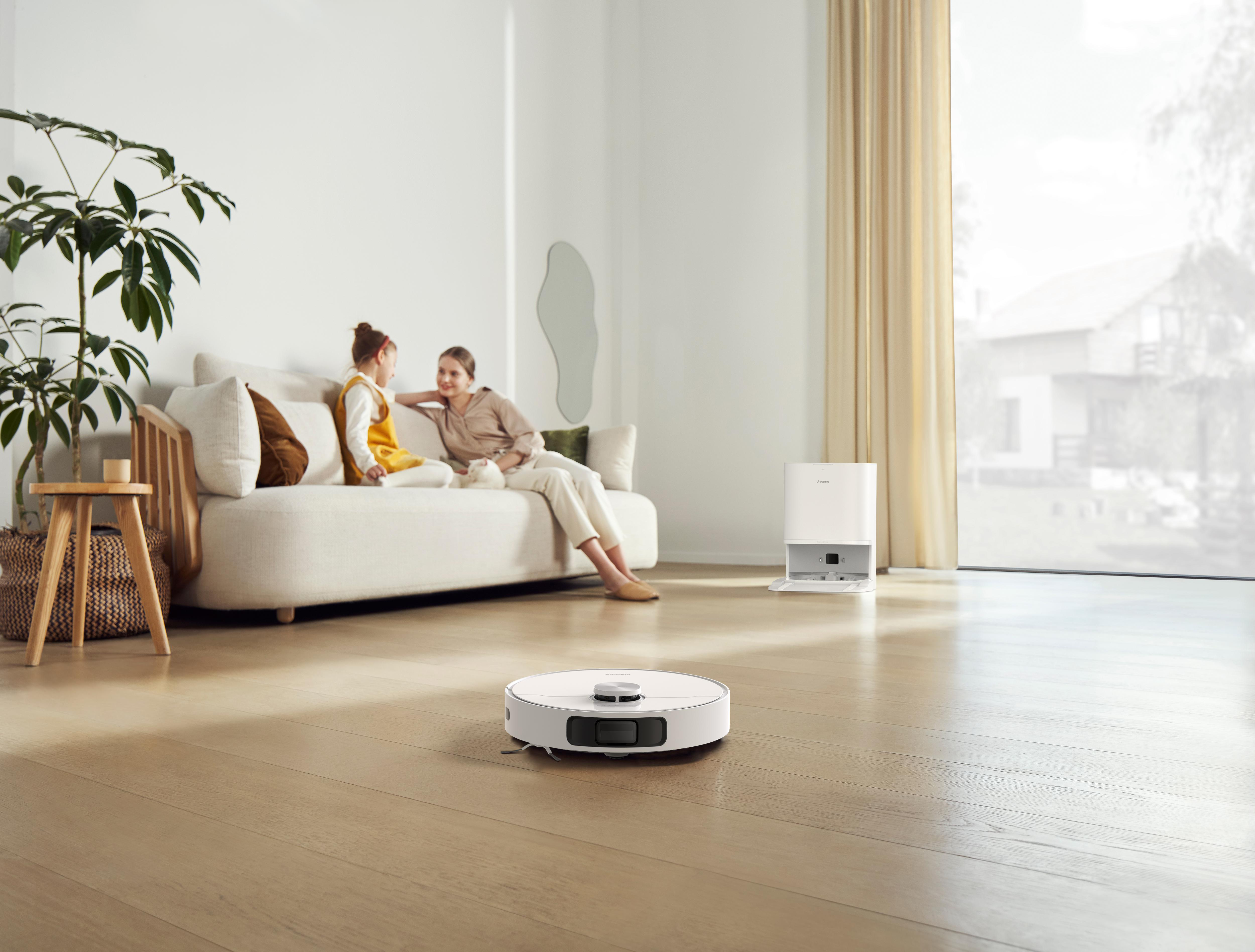 Ein Wohnzimmer mit Personen, weißem Sofa und Roboterstaubsauger auf dem Boden.