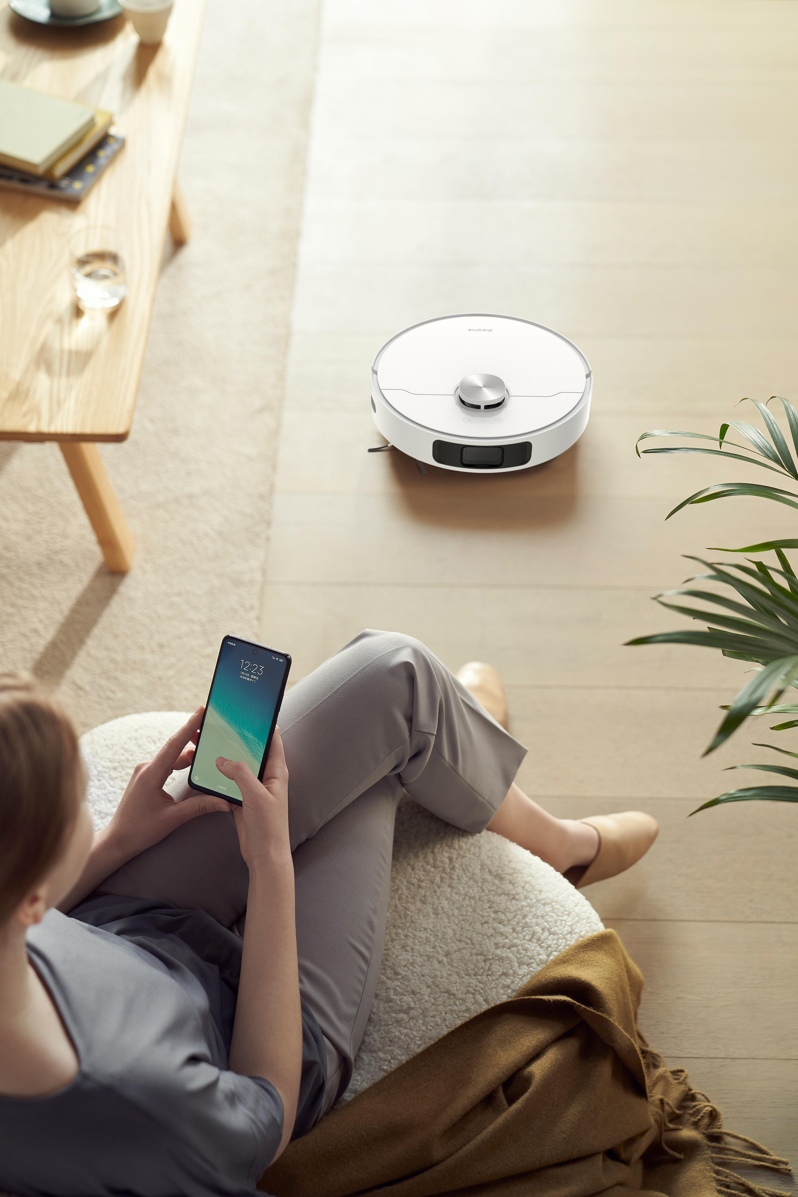 Eine Person, die ein Smartphone benutzt und einem Roboter-Staubsauger beim Reinigen des Bodens zusieht.