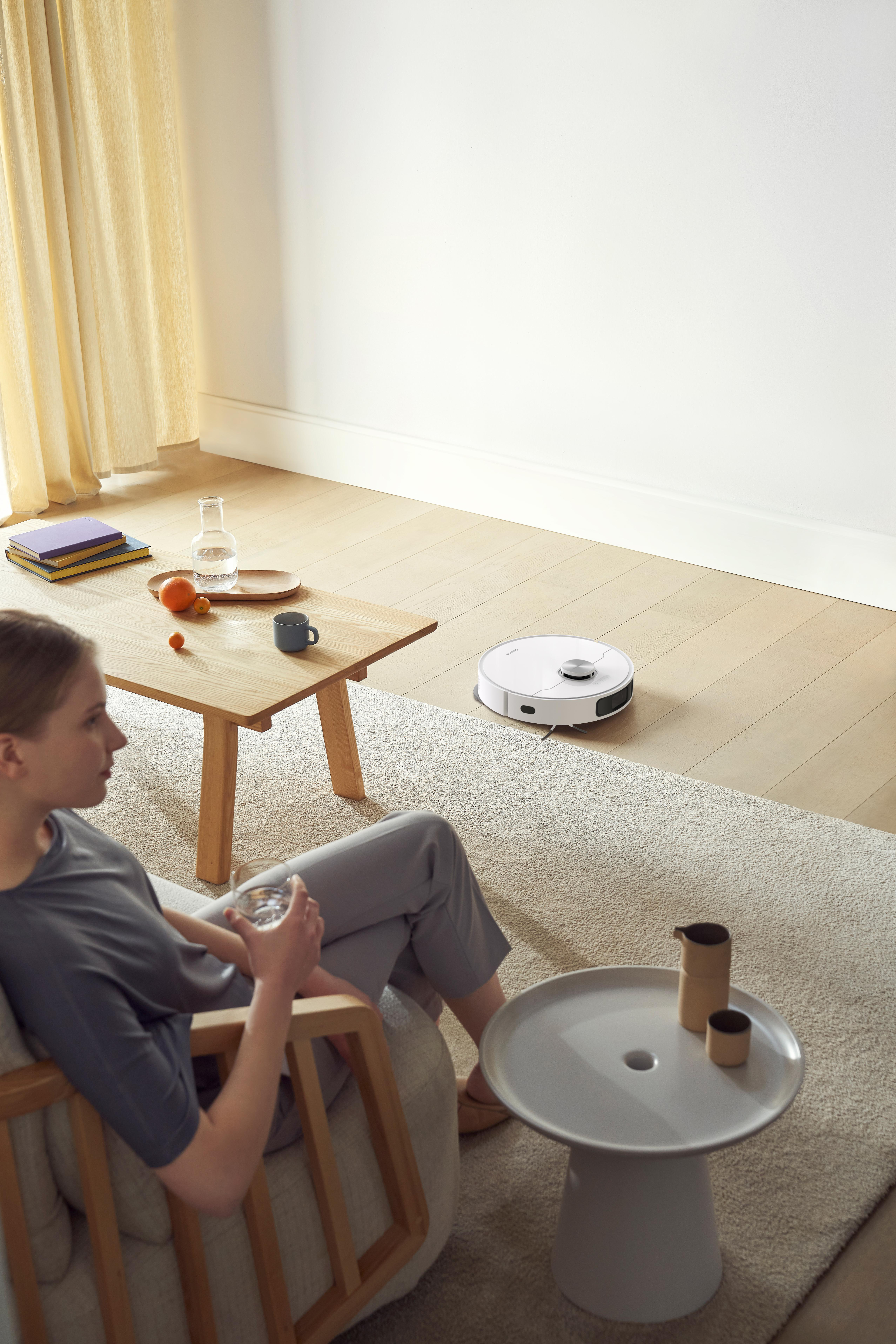 Eine Frau sitzt drinnen und beobachtet einen Roboter-Staubsauger auf einem hellen Teppich. Tisch und Vase sind in der Nähe.