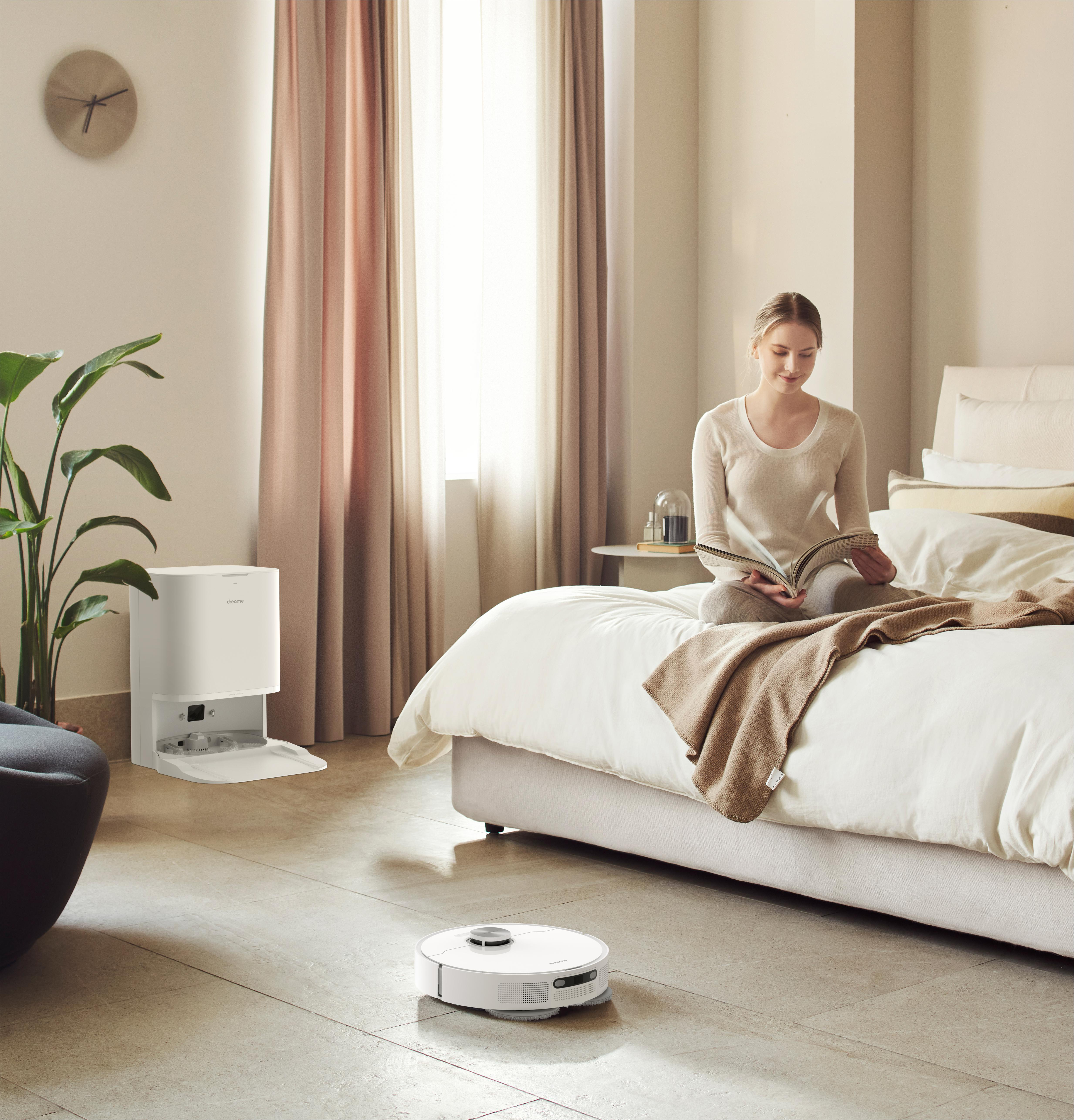 Eine Frau liest im Bett. Ein Reinigungsroboter befindet sich im Vordergrund. Eine Vase und Vorhänge im Hintergrund.