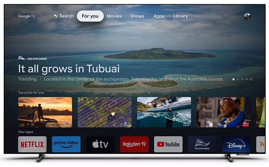 Ekran telewizora pokazujący interfejs Google TV z motywem podróżniczym i ikonami aplikacji u dołu.