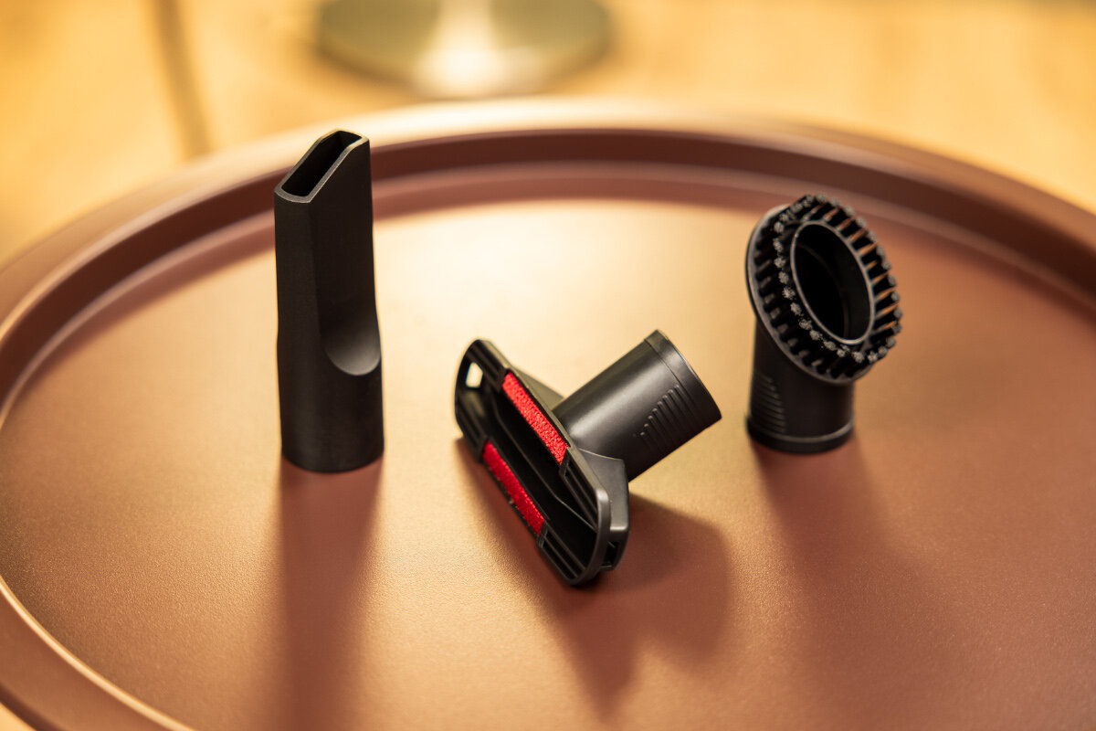 Akcesoria do odkurzacza ułożone na brązowej powierzchni, w tym ssawka szczelinowa i głowica szczotkowa.