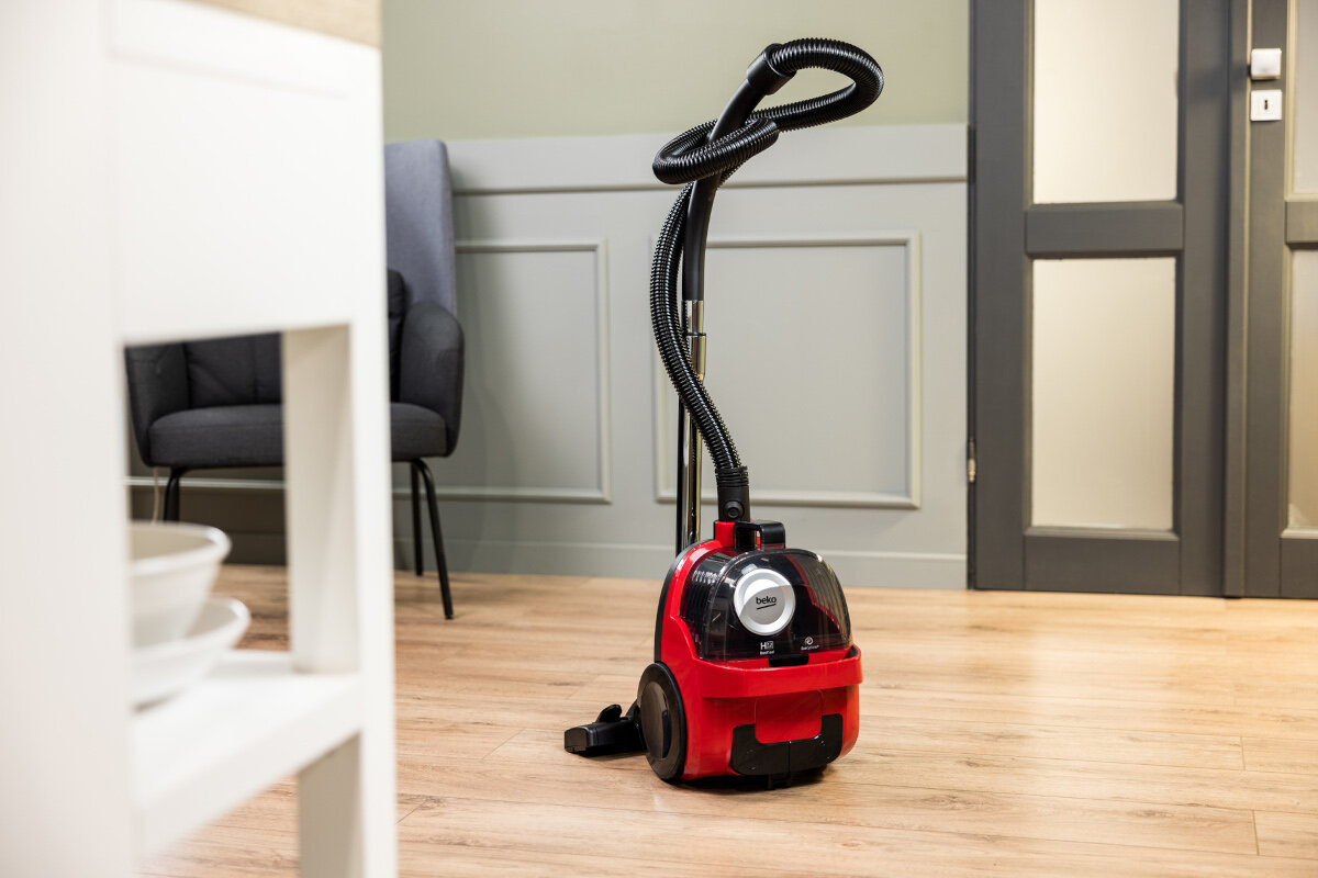 Czerwony odkurzacz stoi na drewnianej podłodze przed ścianą i drzwiami.