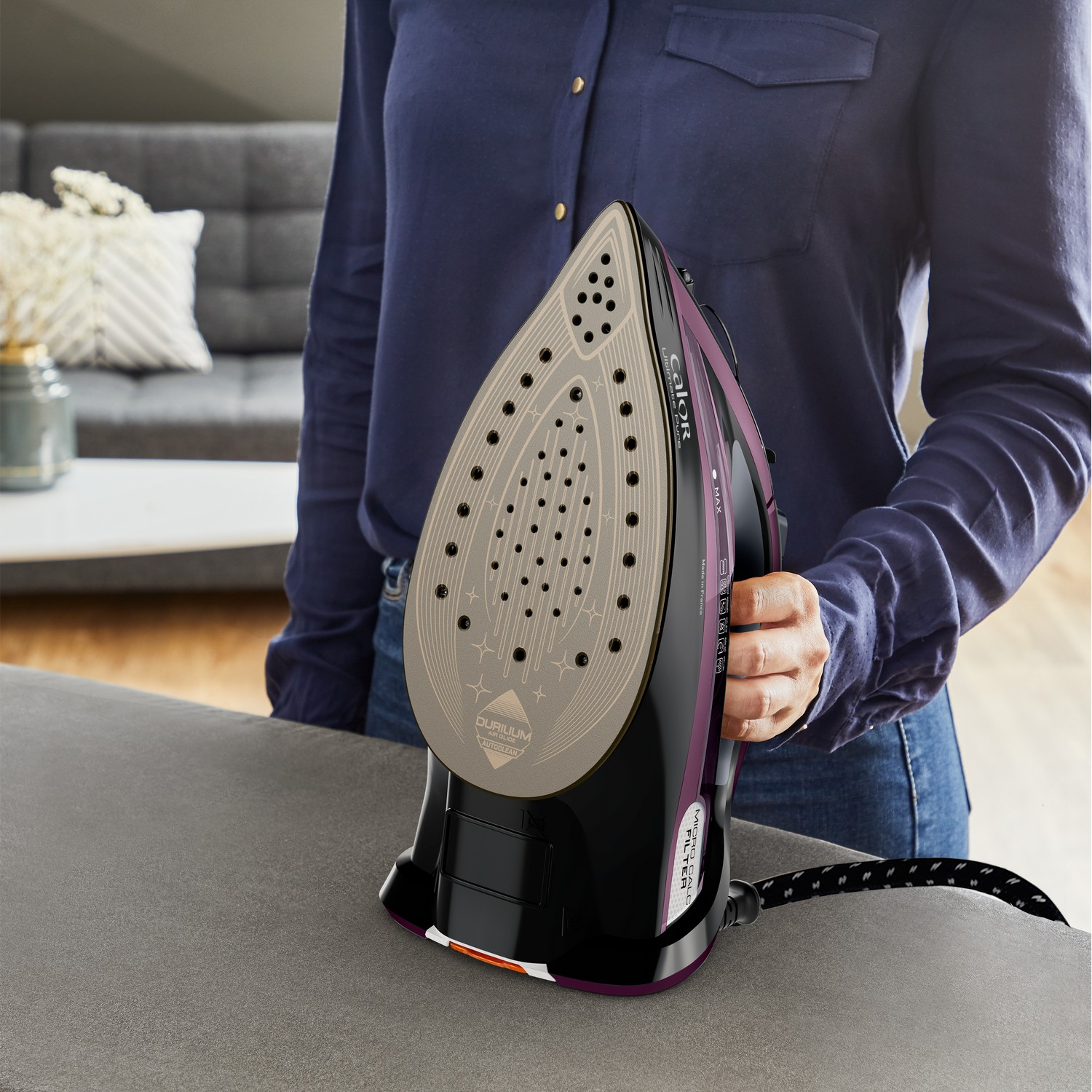 Person holding a black and purple iron over a gray ironing board.