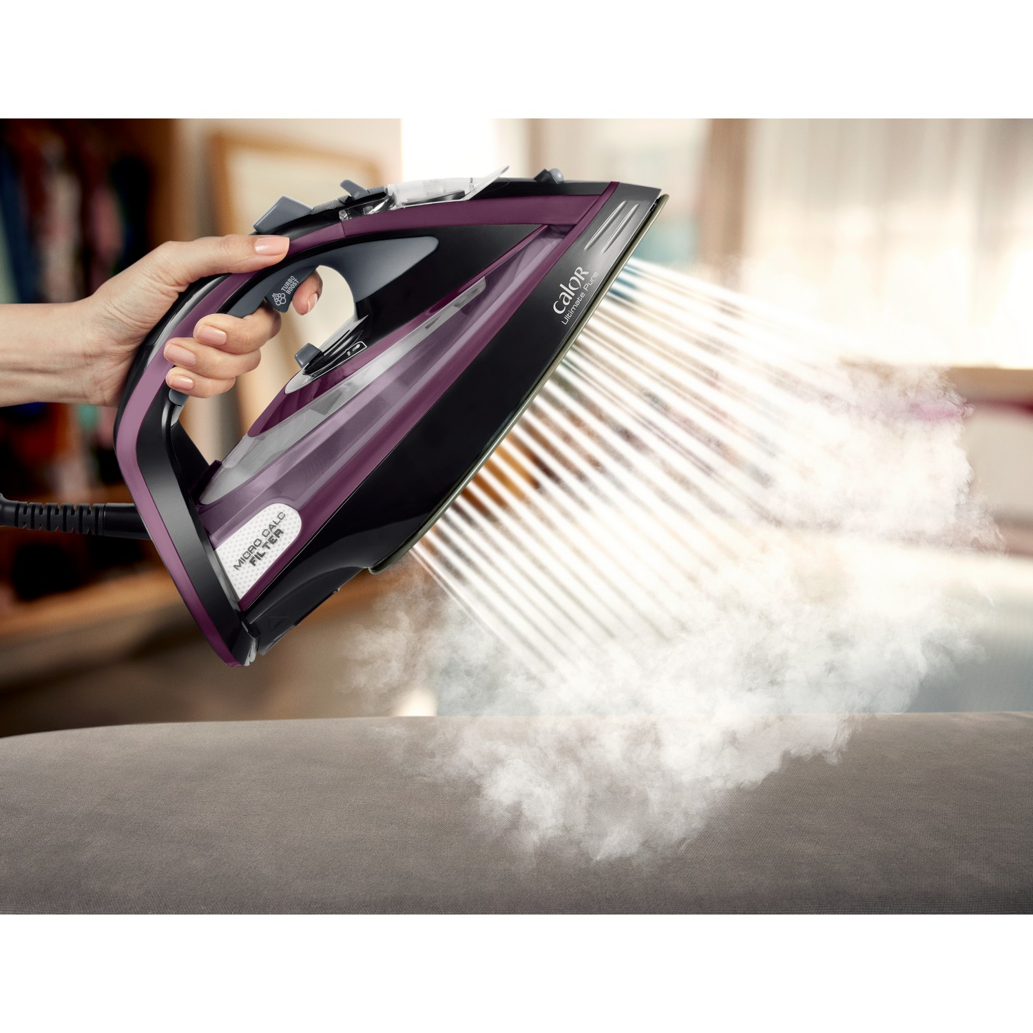 A person's hand holds a purple iron, steam emerging, over a grey sofa. Background blurred, room with clothes and window.