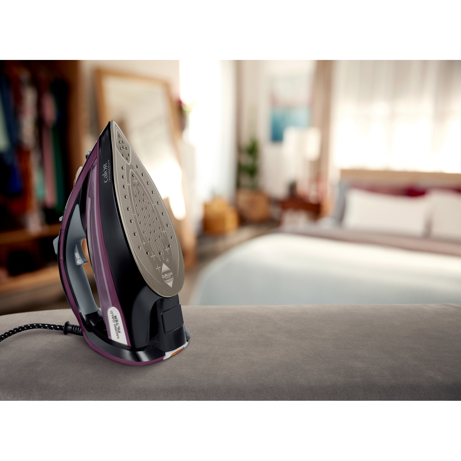 An iron on a grey surface in a room with a mirror and clothes.