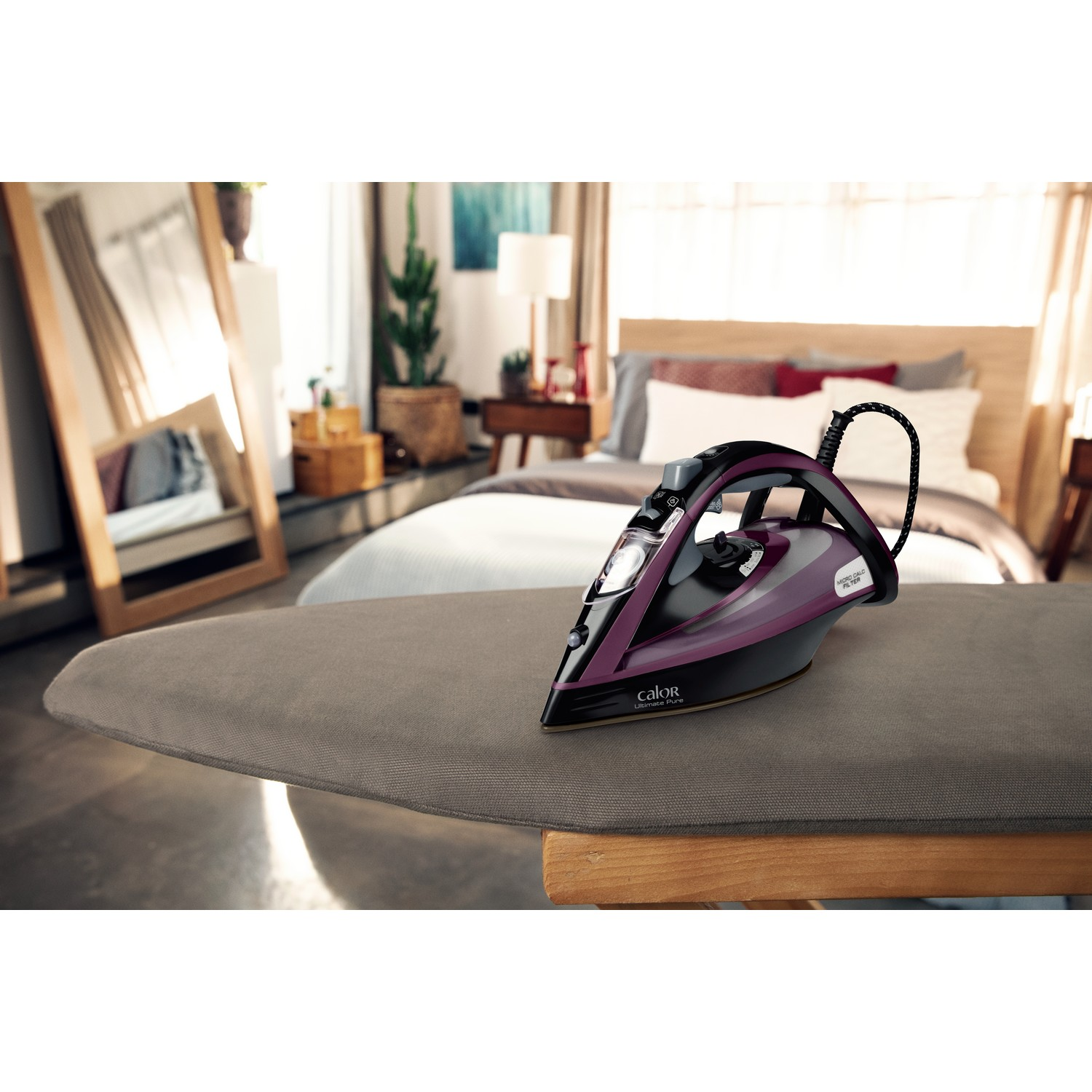 Black and purple Calor iron on an ironing board with mirror and bed background.