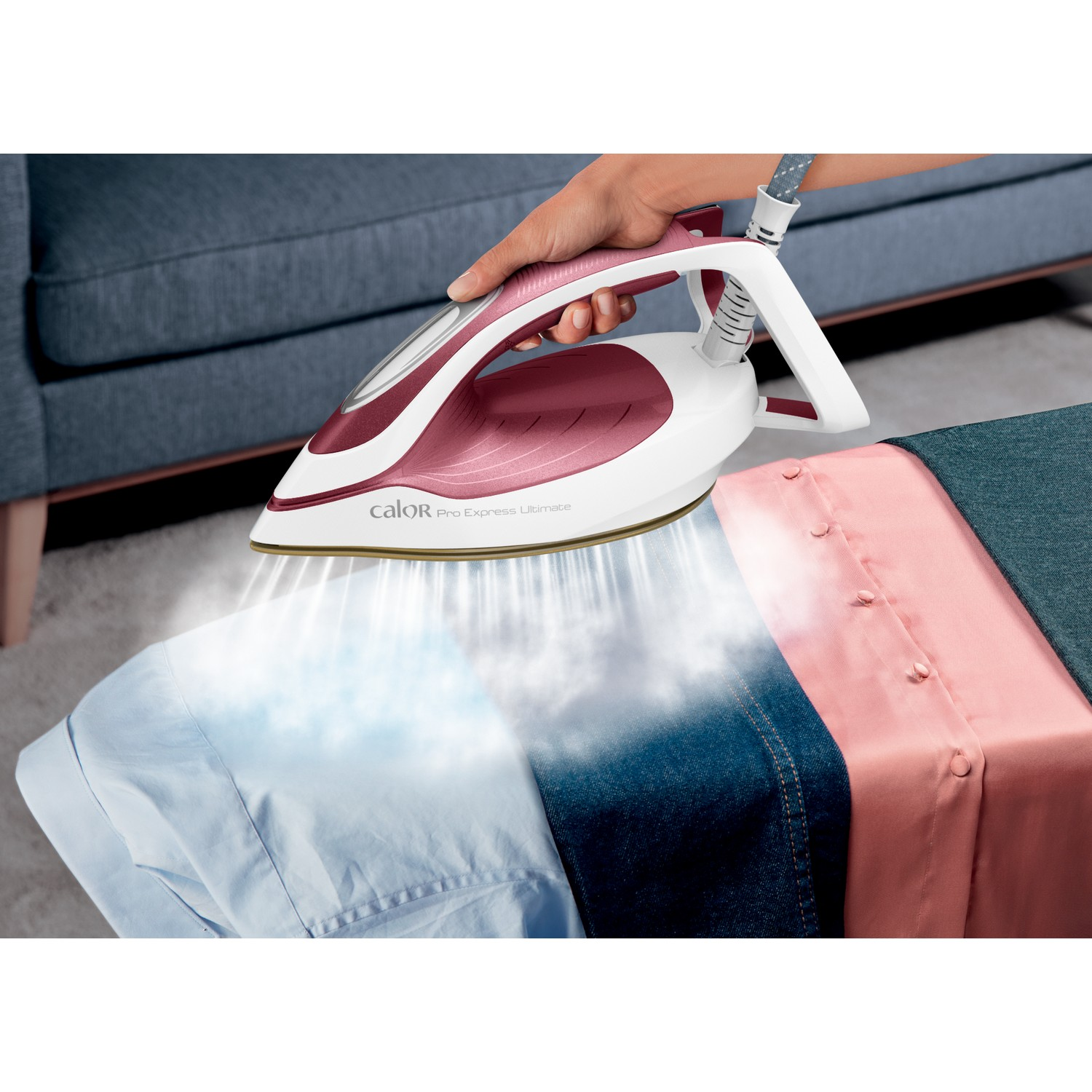 A hand ironing a shirt with a pink and white iron, steam visible, with blue and pink fabrics.