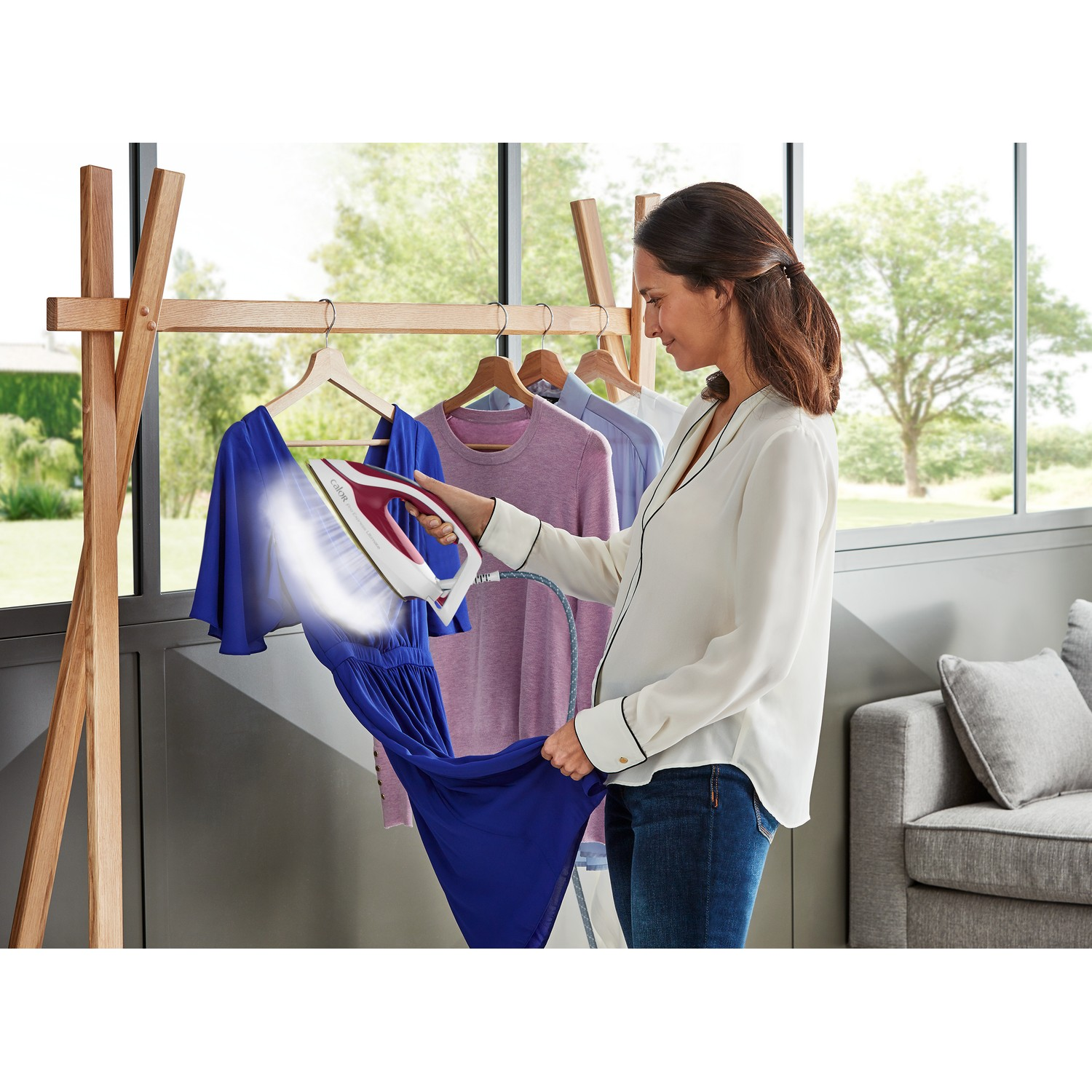 A woman ironing a blue dress on a hanger. Clothes rack in the background. Window and a couch.