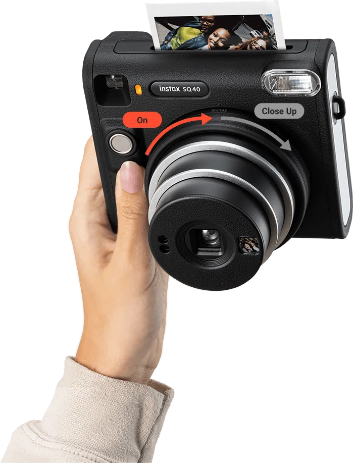 Appareil photo Instax noir avec éjection de photos. Une main tient l'appareil. Boutons rouges et blancs visibles.