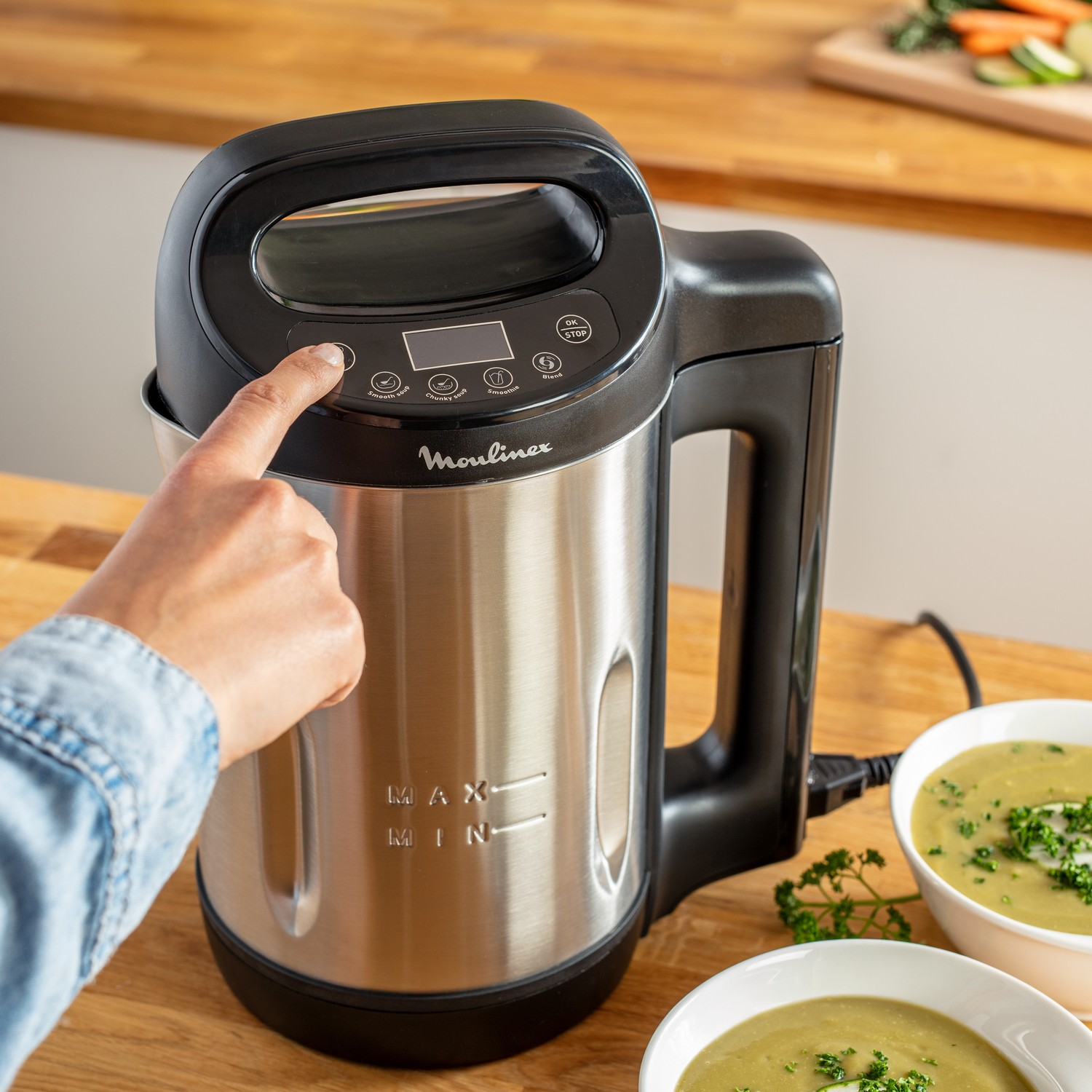 A person is pressing a button on a silver and black soup maker machine.