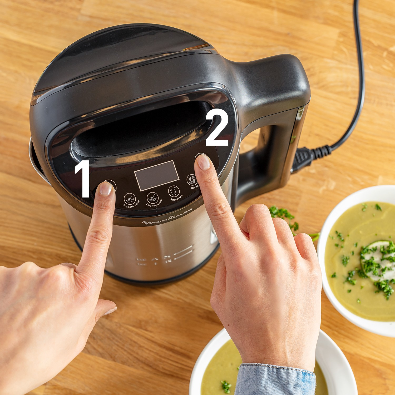 Hands pressing buttons on a black and silver food processor, with soup bowls.