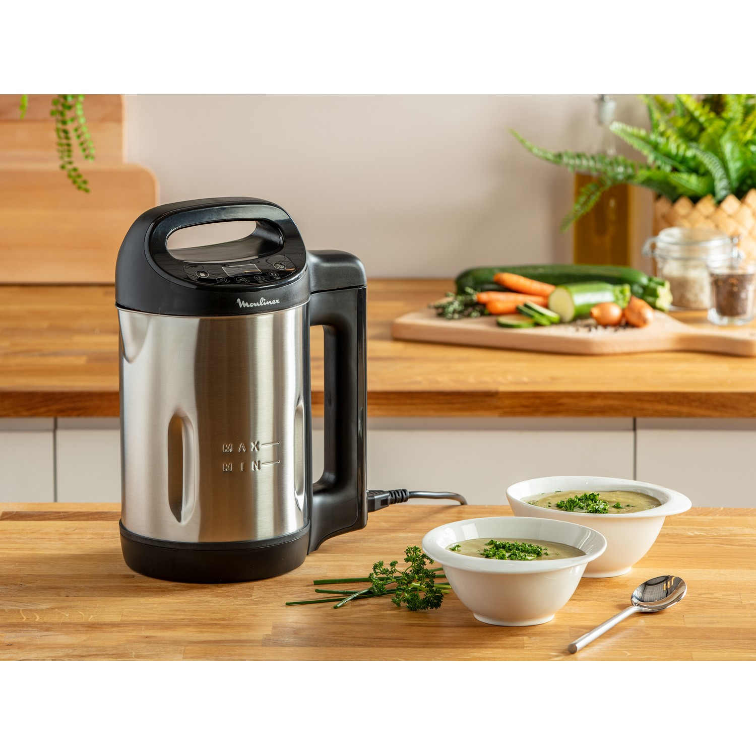 Stainless steel soup maker with handle. Two bowls of soup and vegetables on wooden table.