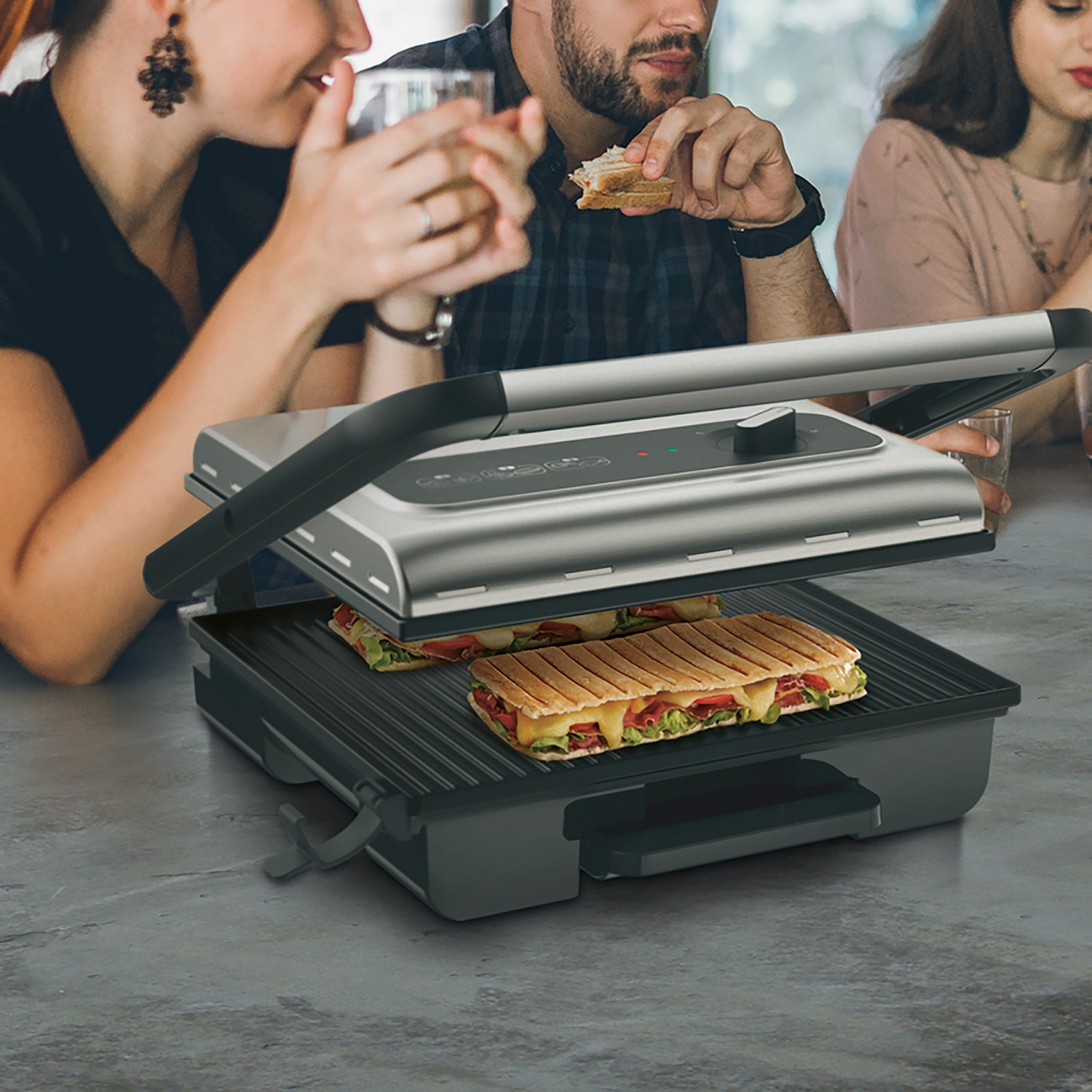 Un grill à panini avec deux sandwichs. Des personnes sont assises à une table en arrière-plan.