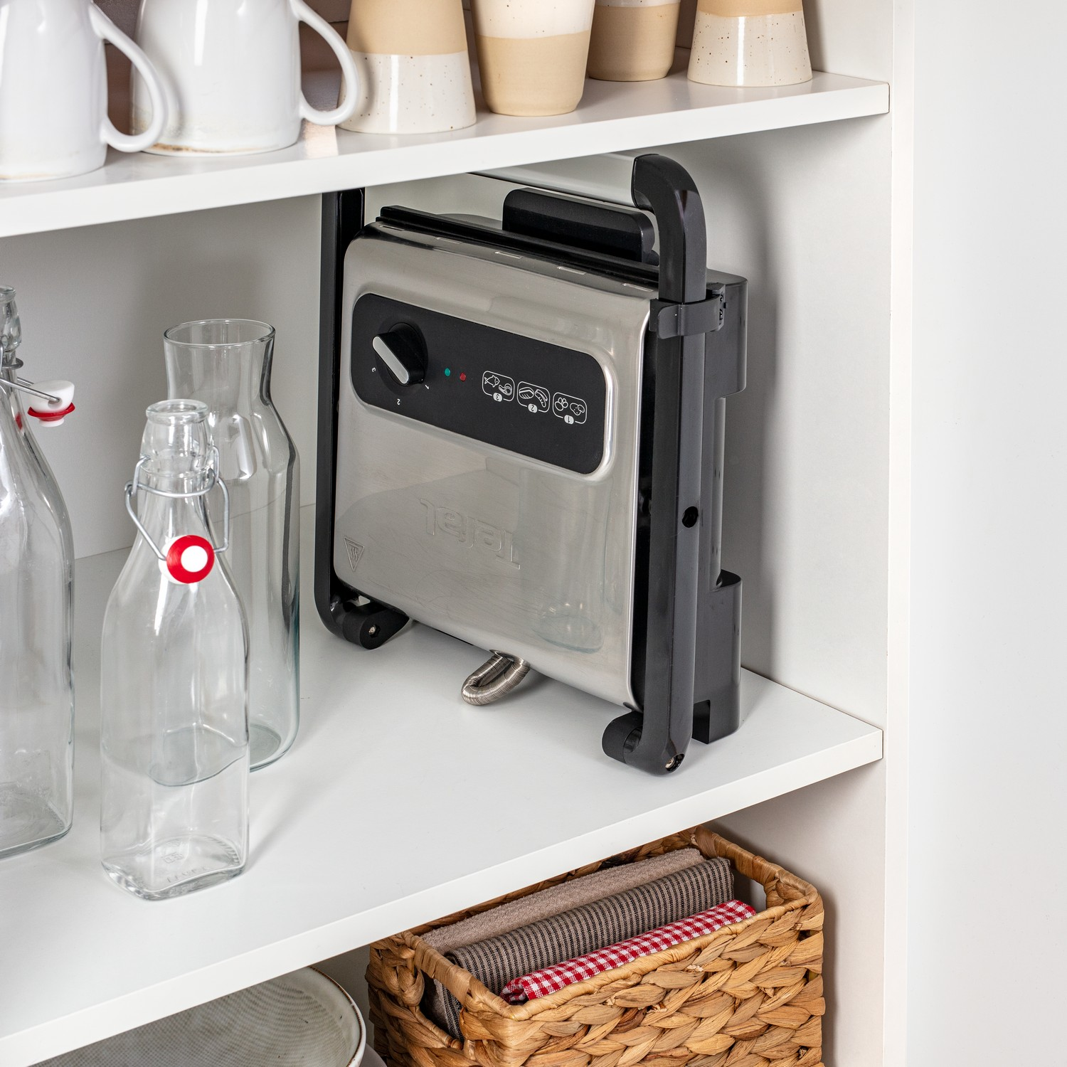 Un grill Tefal argenté à l'intérieur d'un placard blanc. Bouteilles et bocaux au-dessus et en dessous.