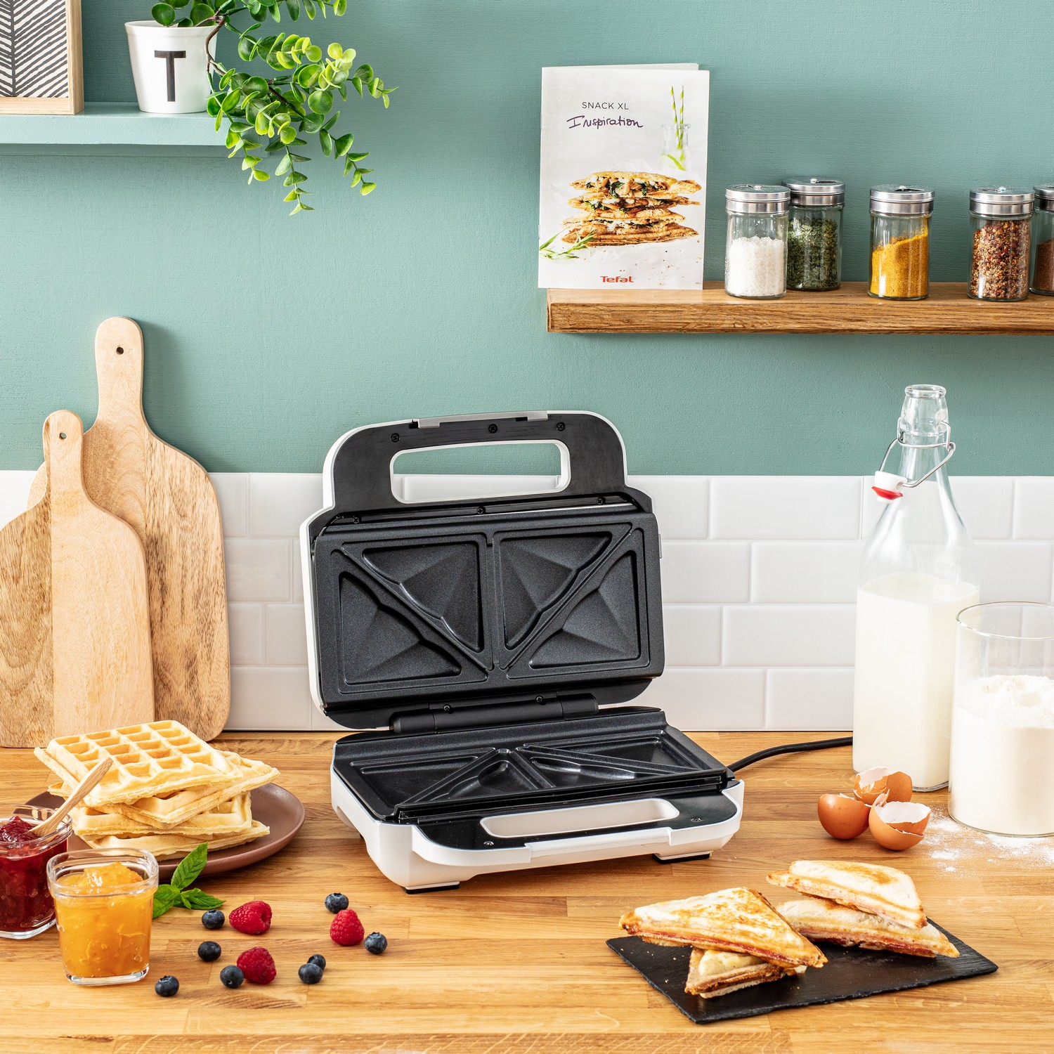 Sandwich maker on a wood table. Waffles, jam, fruit, and milk are on the table.