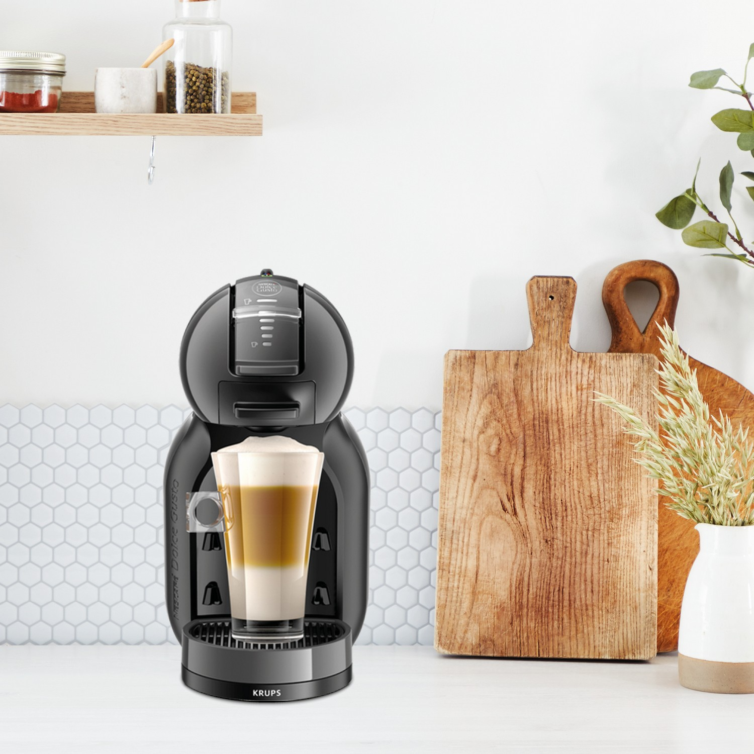 Coffee machine on a white counter. A latte is in a glass.  A wooden cutting board and vase are in the background.