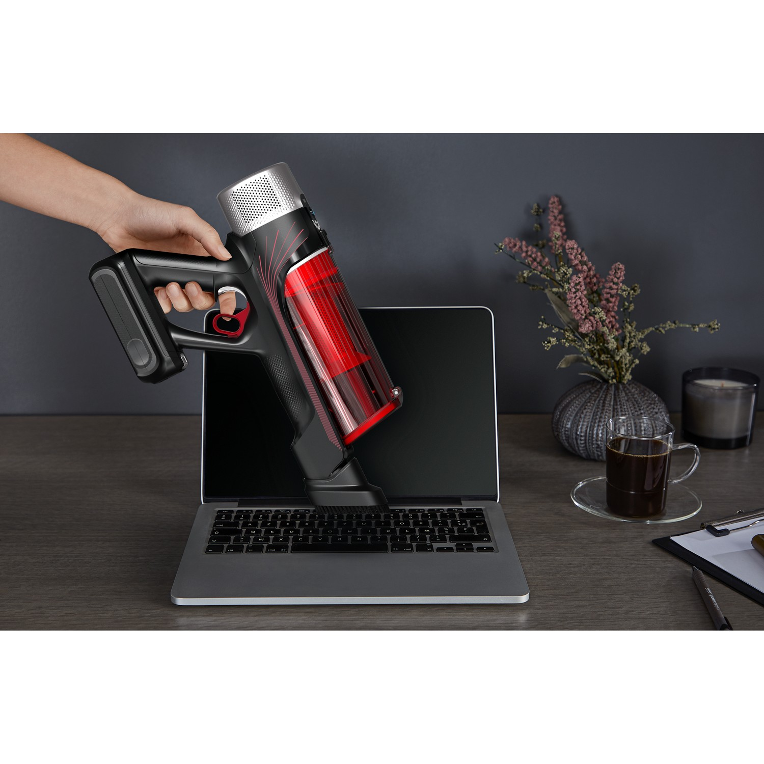 Een persoon houdt een stofzuiger boven een laptop. Een kopje koffie en bloemen staan op tafel.