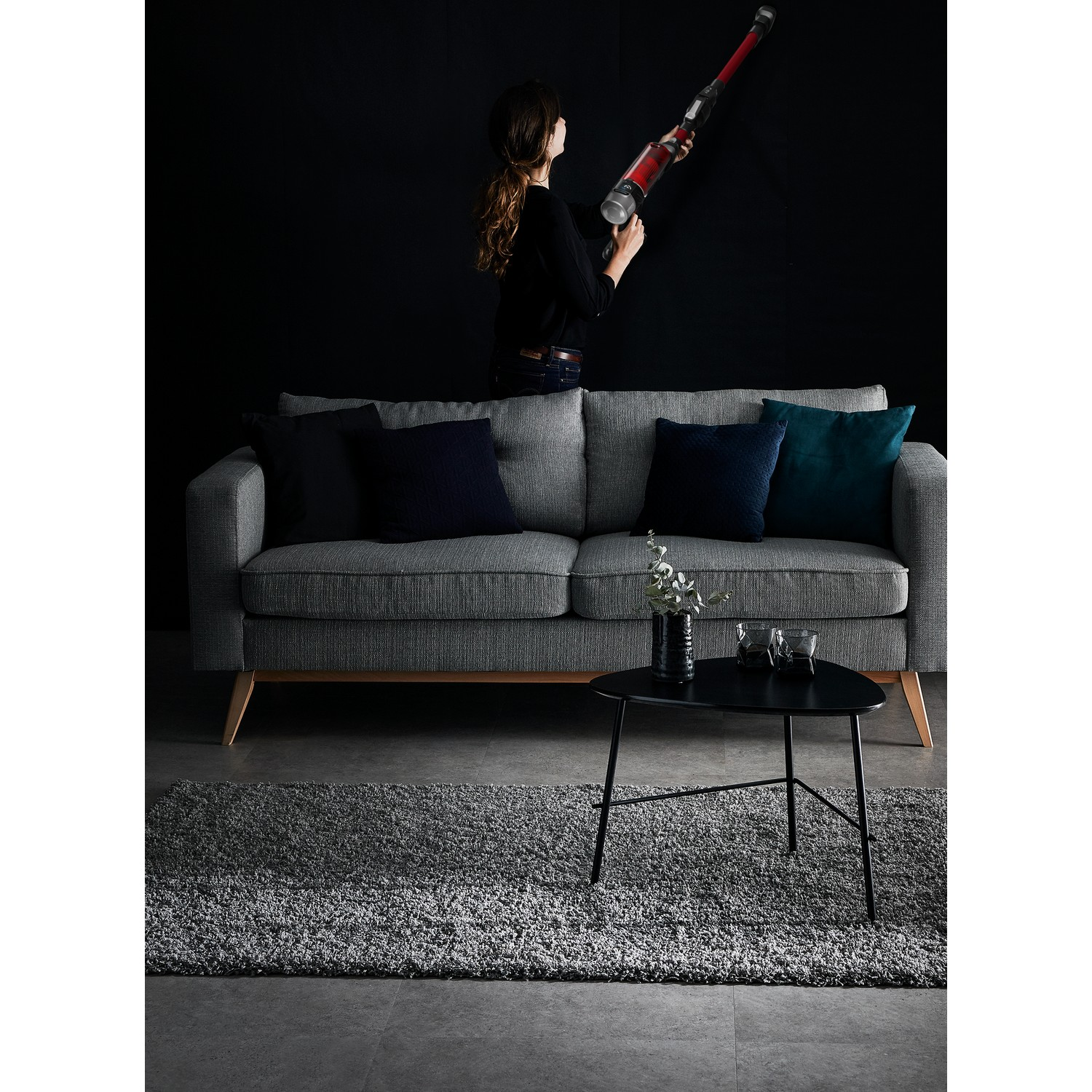 Een vrouw stofzuigt een zwarte muur. Een grijze bank, tafel en tapijt staan vooraan. Twee glazen en een vaas staan op de tafel.