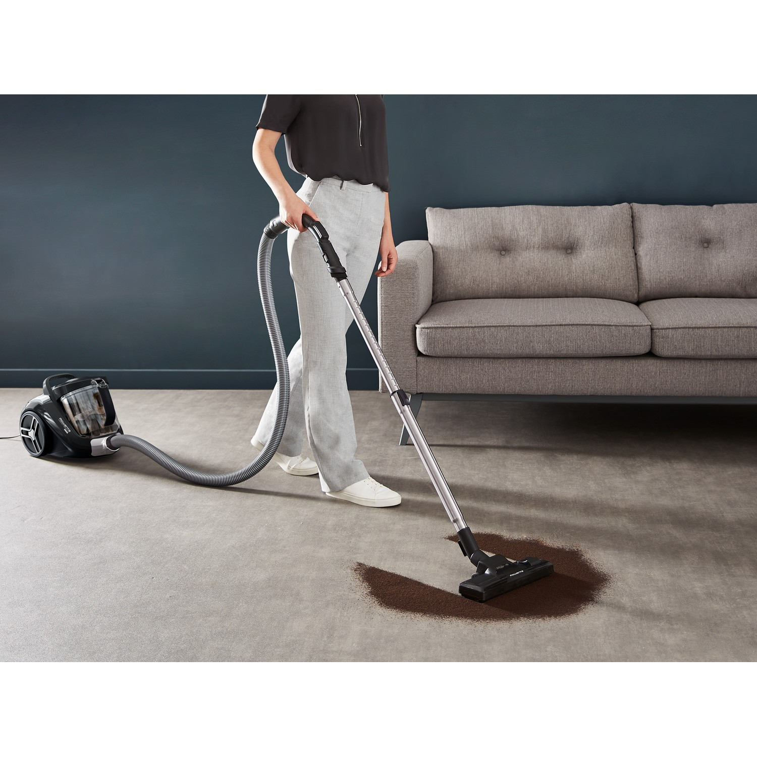 A person vacuuming a carpet with a black vacuum cleaner, with a sofa in the background.