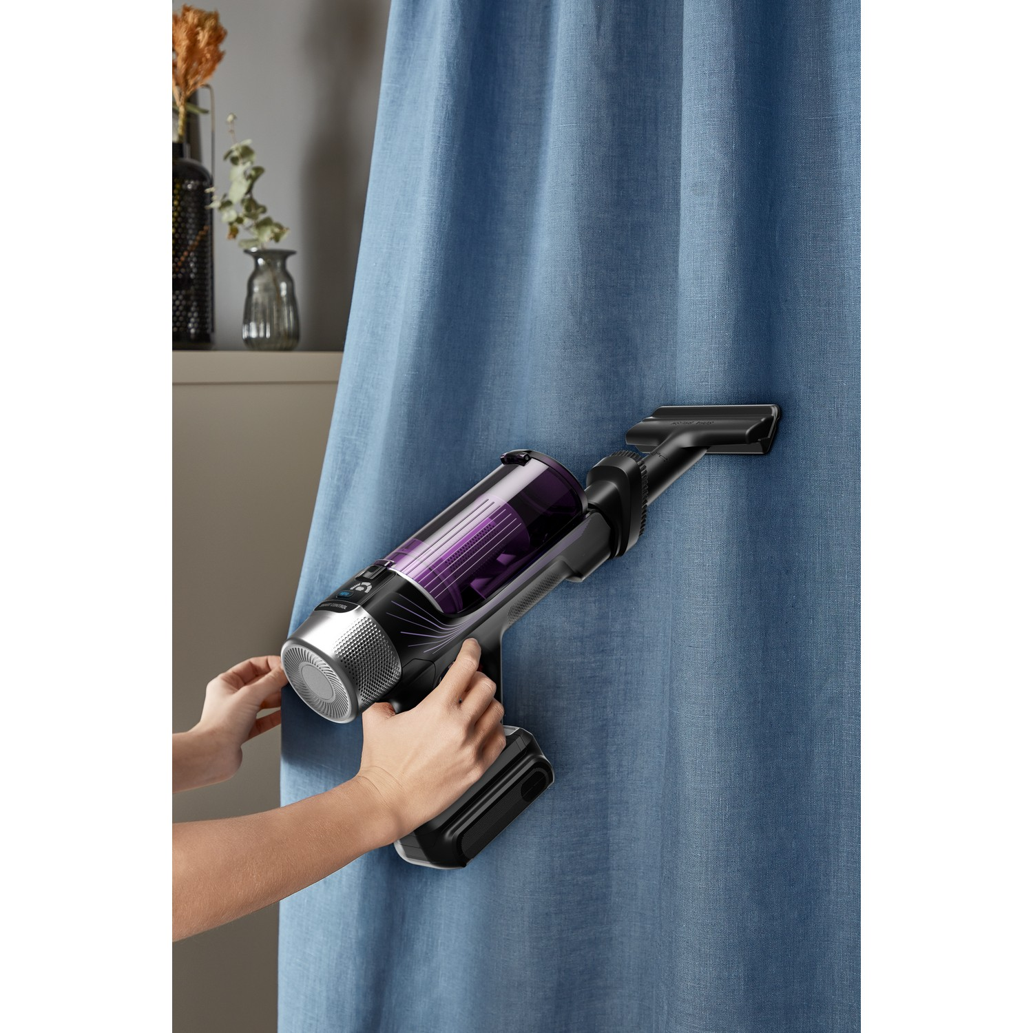 A woman using a vacuum cleaner on a blue curtain. The vacuum is black and purple.