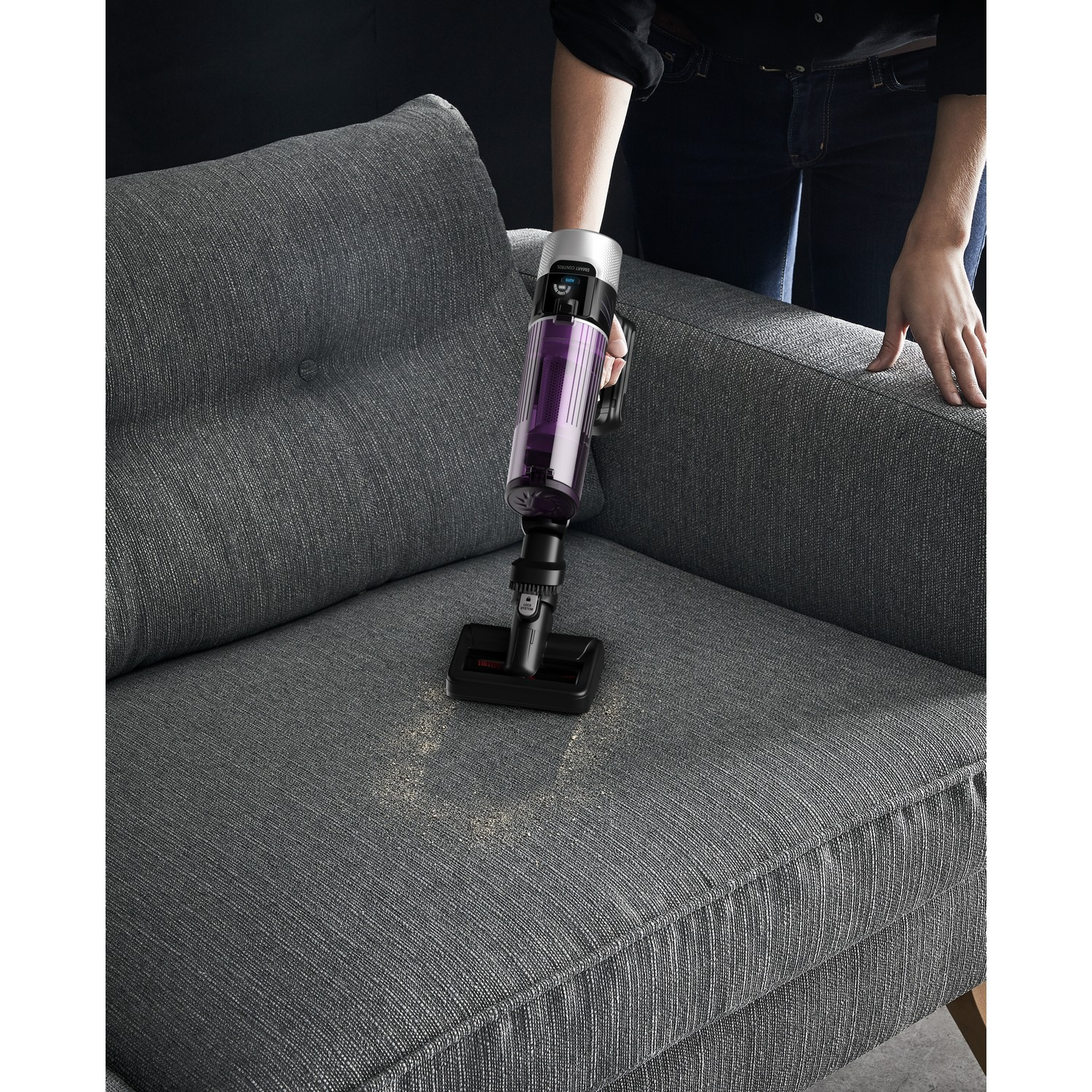 Person vacuuming a gray sofa with a purple and silver handheld vacuum cleaner, with dust visible.