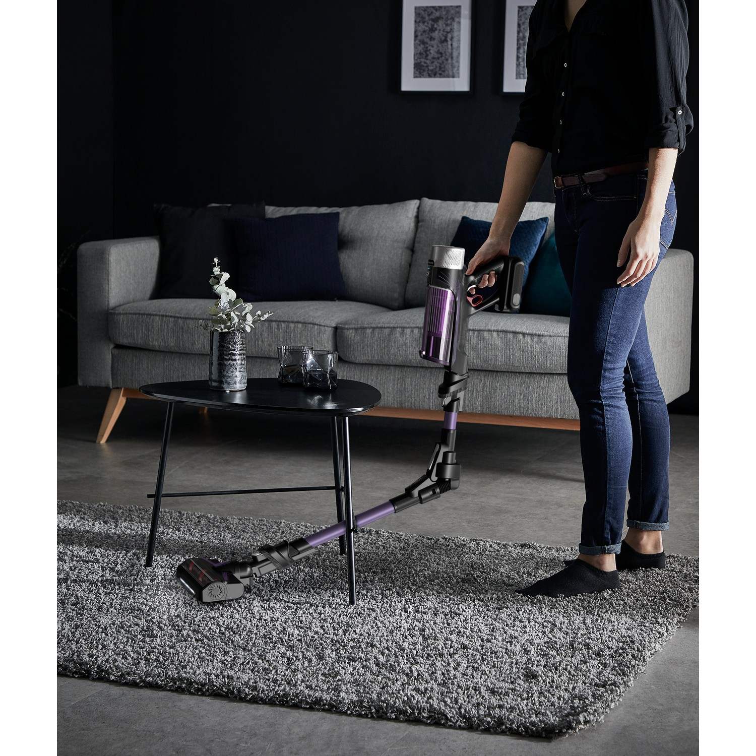 A woman using a purple and black cordless vacuum cleaner on a gray rug near a table.