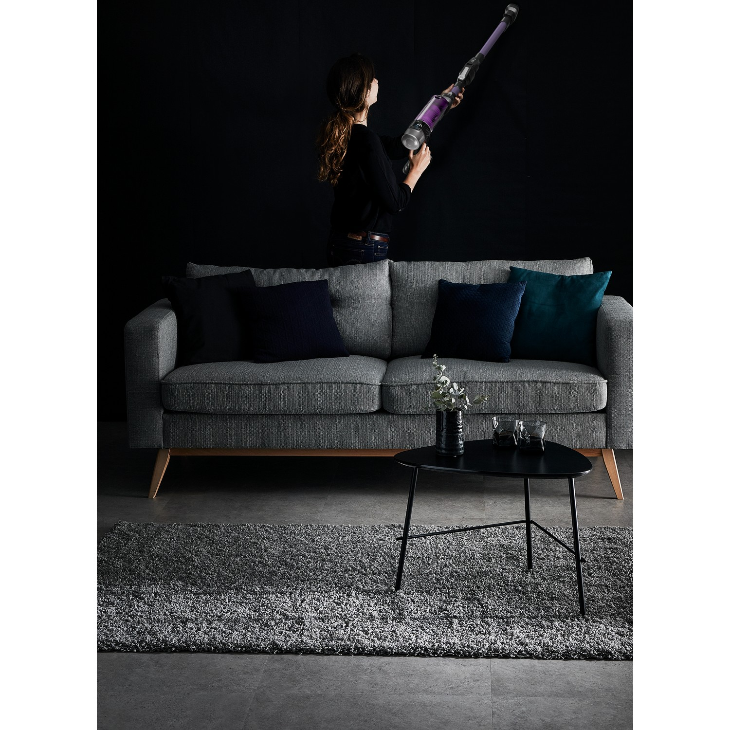 Woman vacuuming. Grey sofa, black table, rug, and black background with the vacuum in use.