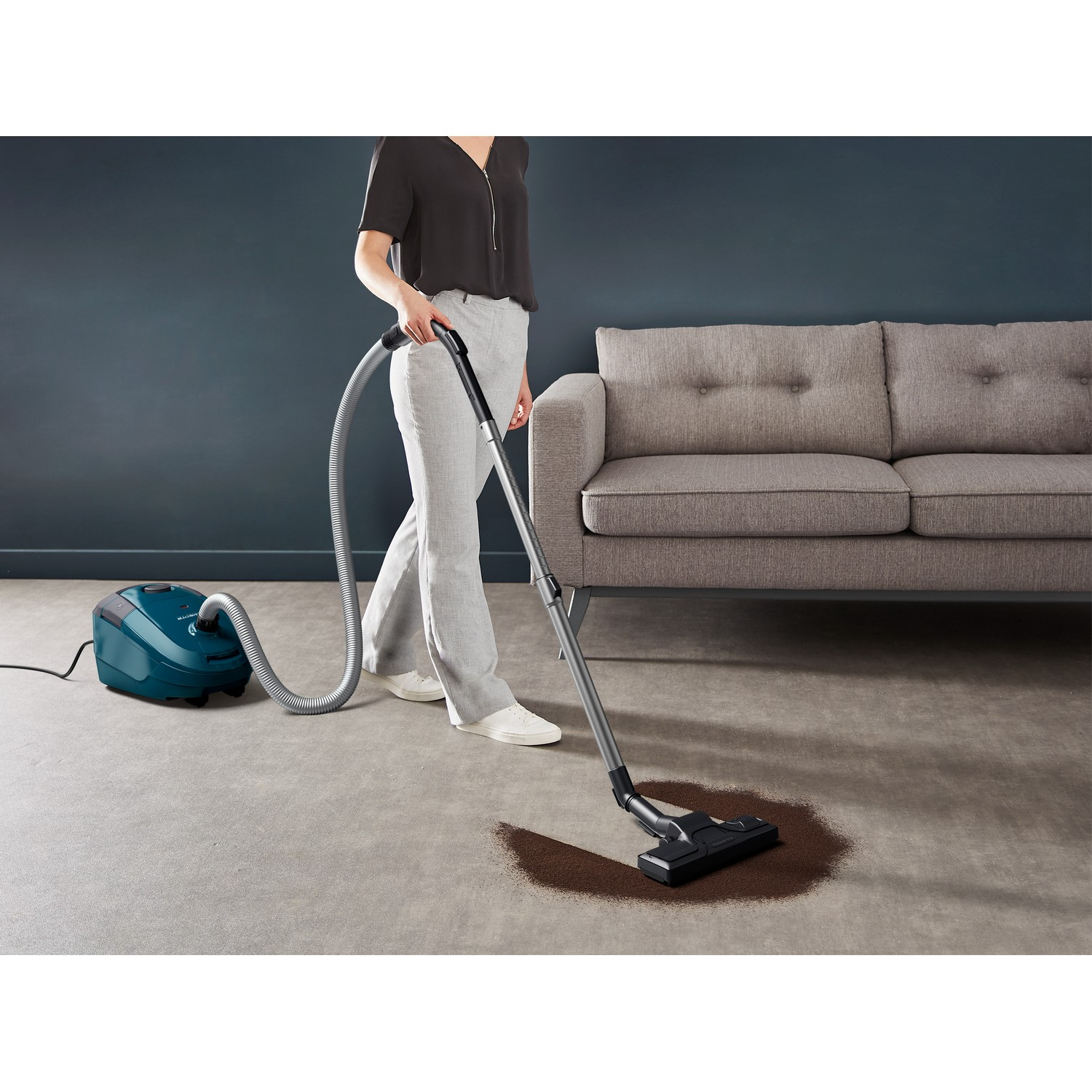 A woman vacuums a carpeted floor. A teal vacuum cleaner is in the background.