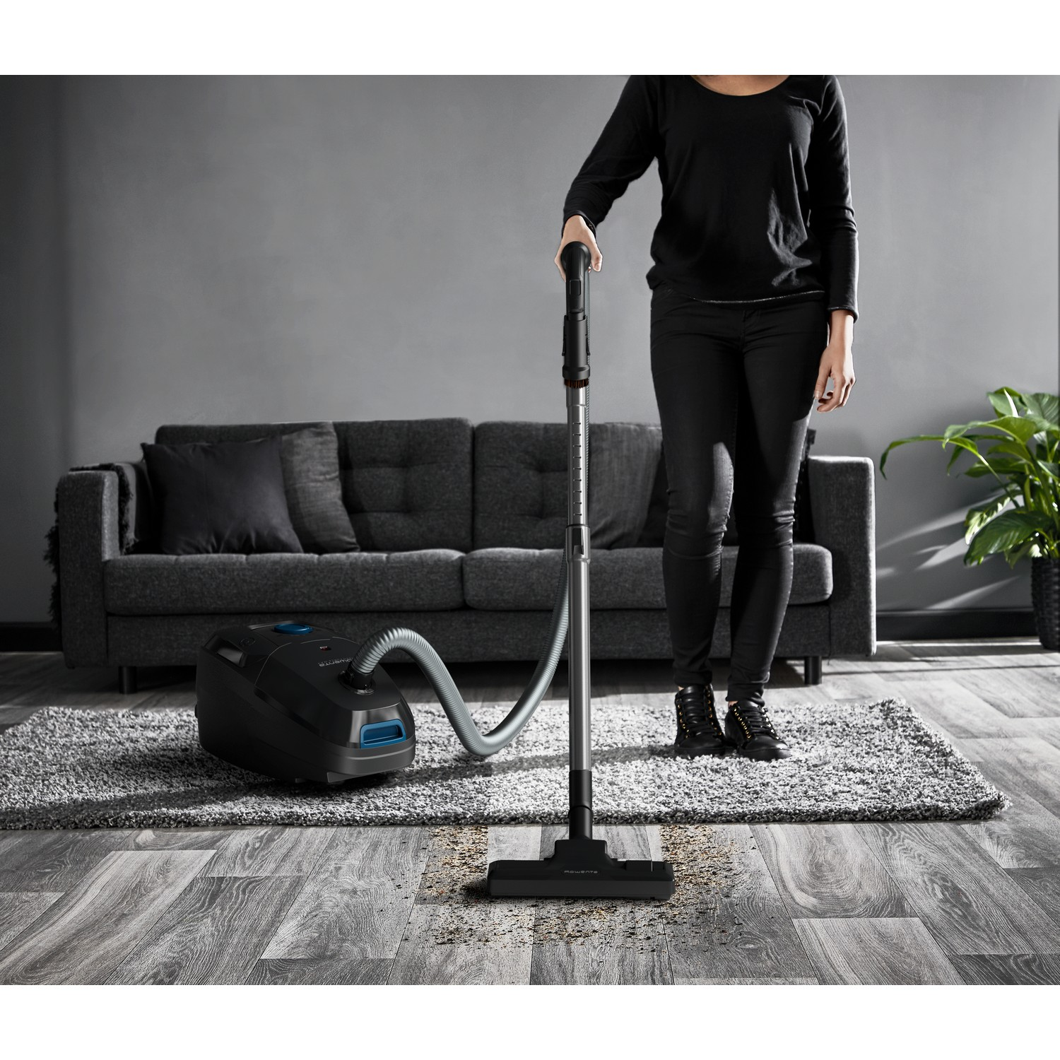Person vacuuming a room. A black vacuum cleaner and a gray sofa are in the background.