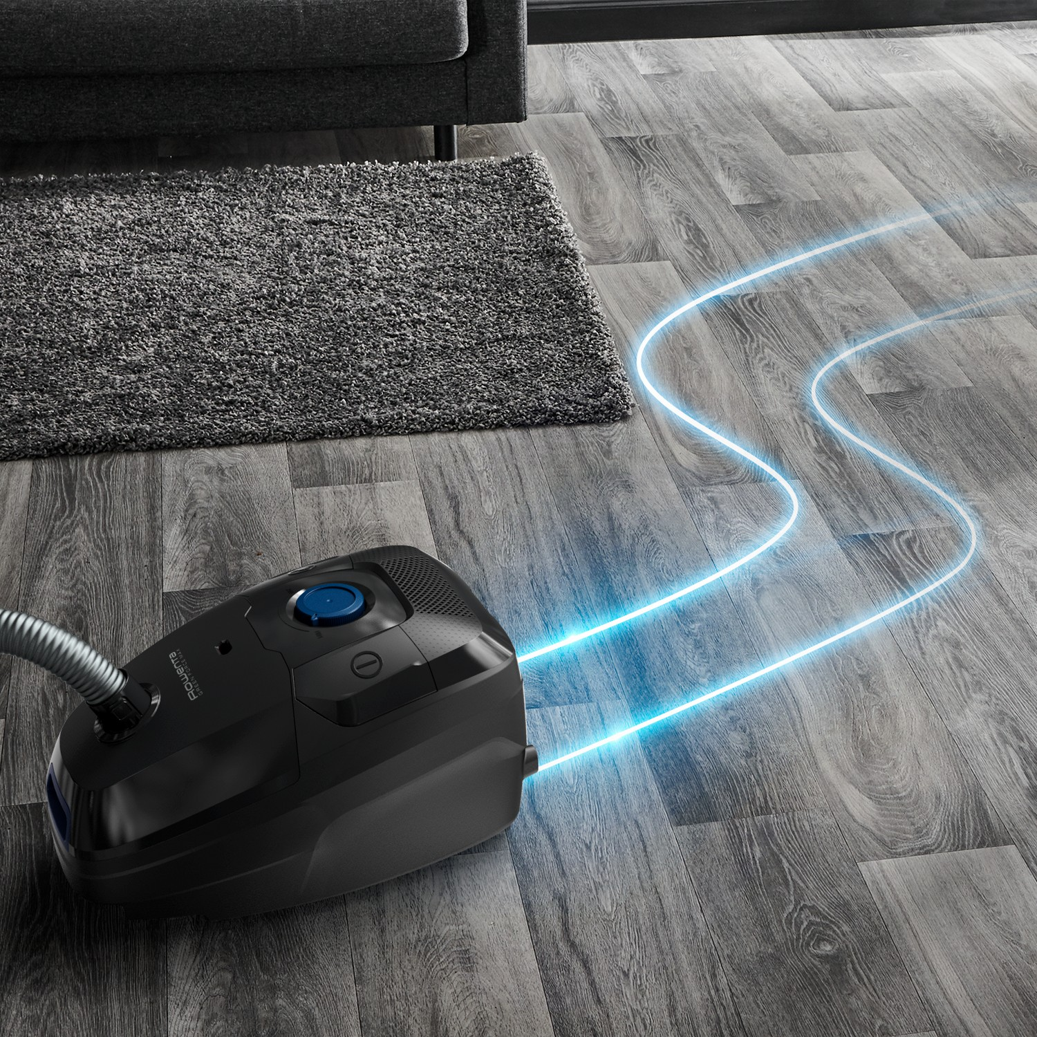 A black vacuum cleaner with blue light trails on a gray wooden floor. A rug is in the background.