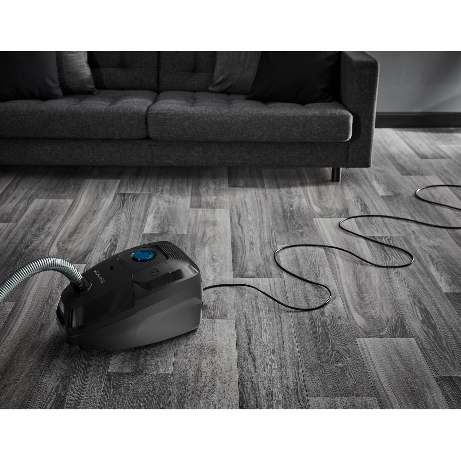 Black vacuum cleaner on gray wooden floor, with a sofa in the background.