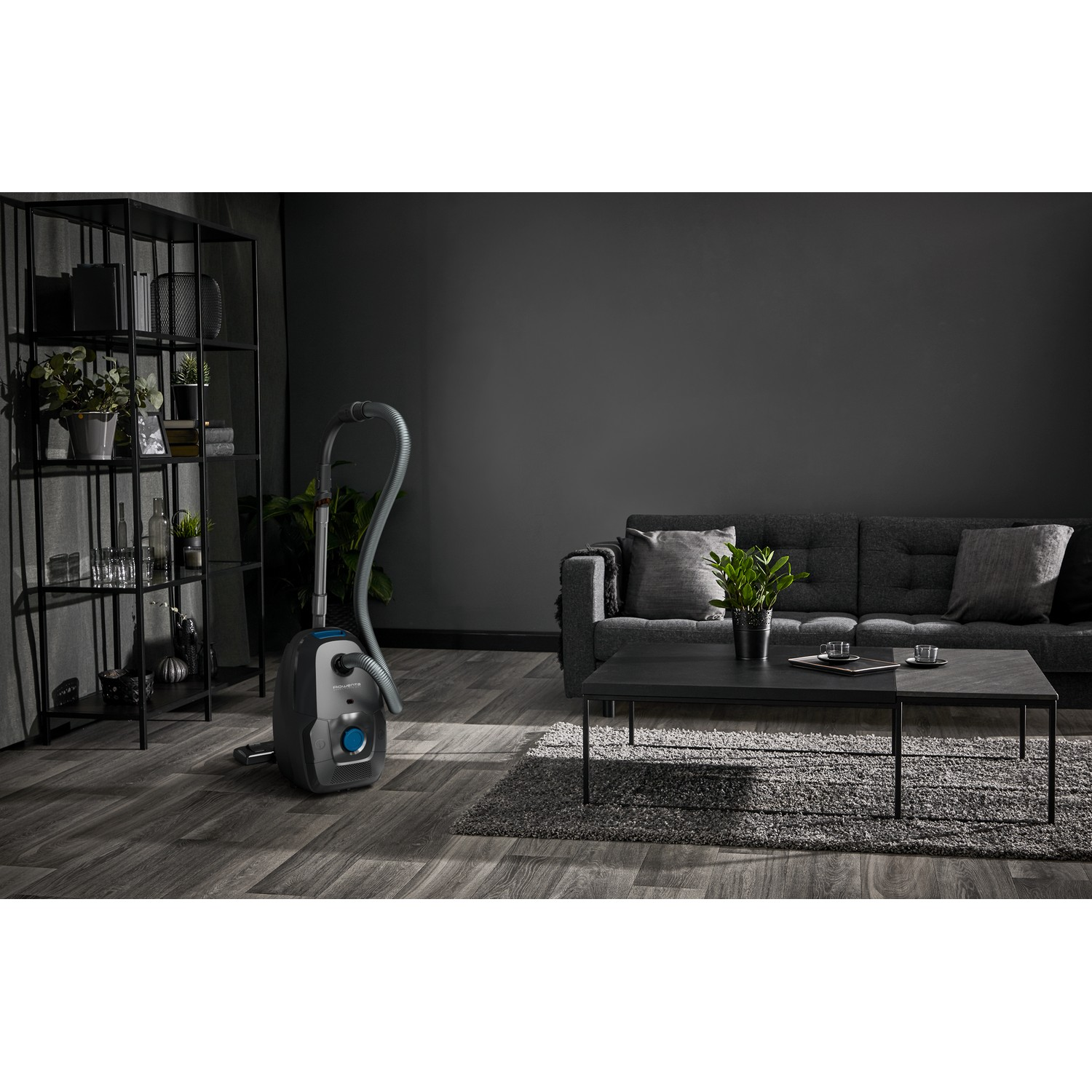 Living room with grey sofa, table, and vacuum cleaner. Dark walls and shelves.
