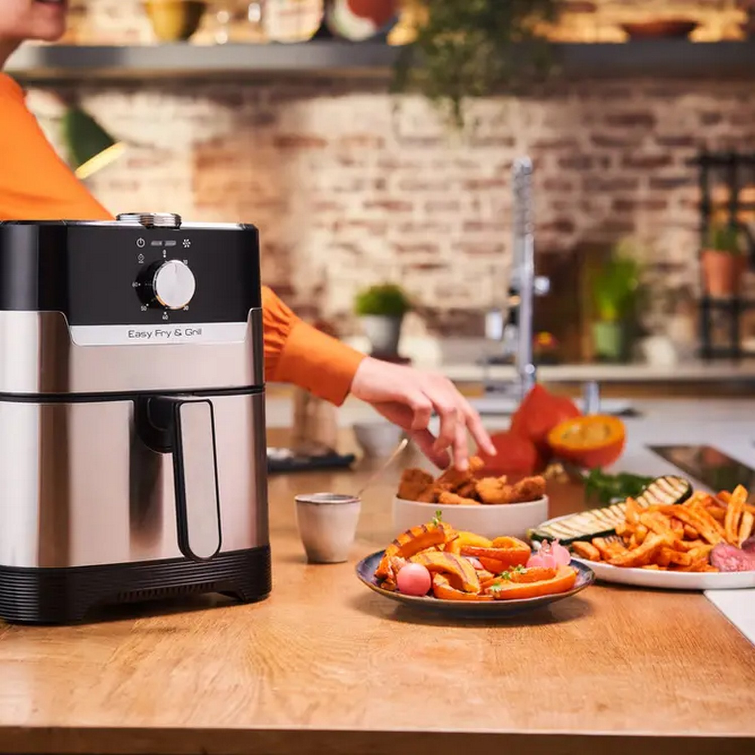 A person in an orange shirt is near a black and silver air fryer. There are food items.