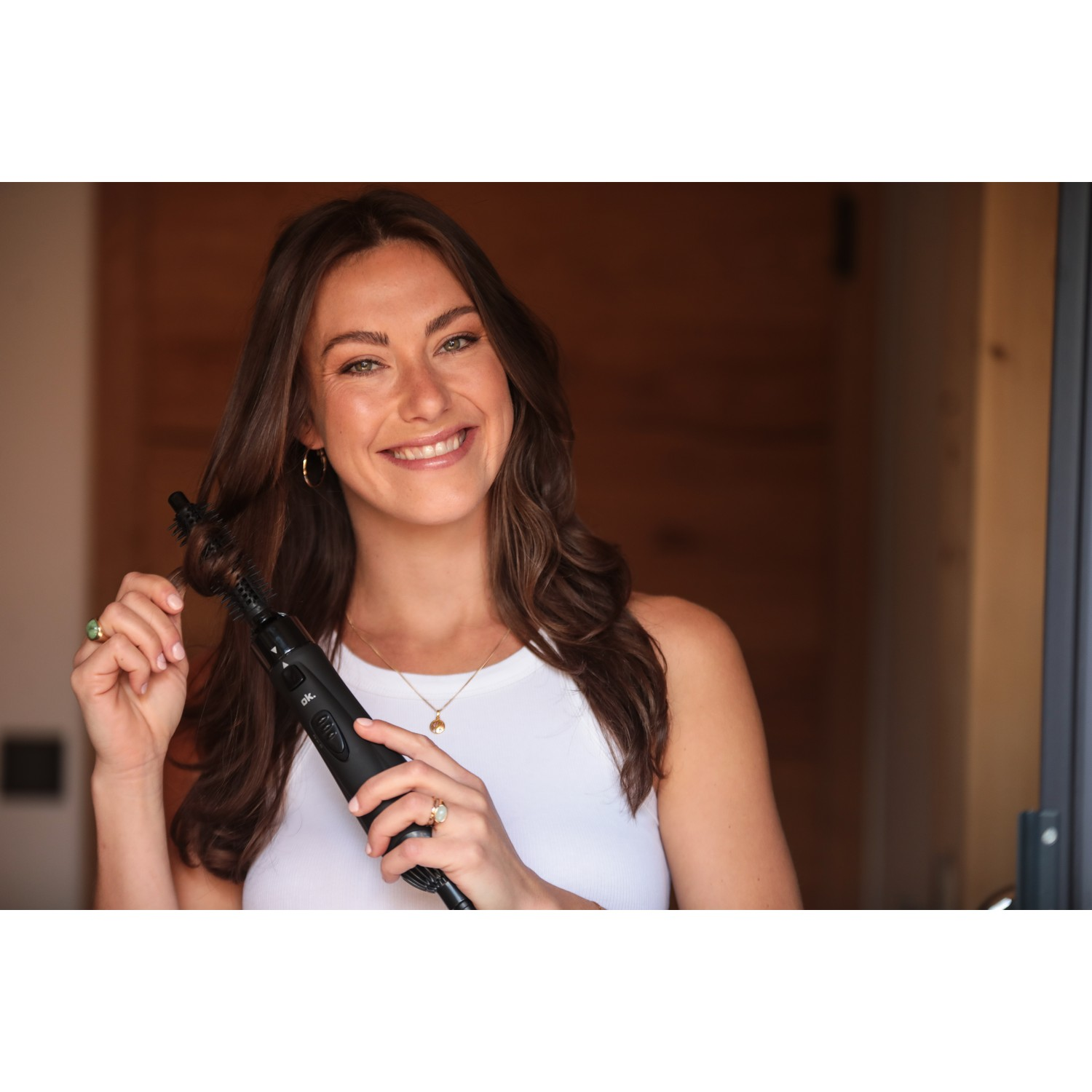 Smiling woman holding a black hair styling tool, wearing a white top. Wooden door in background.