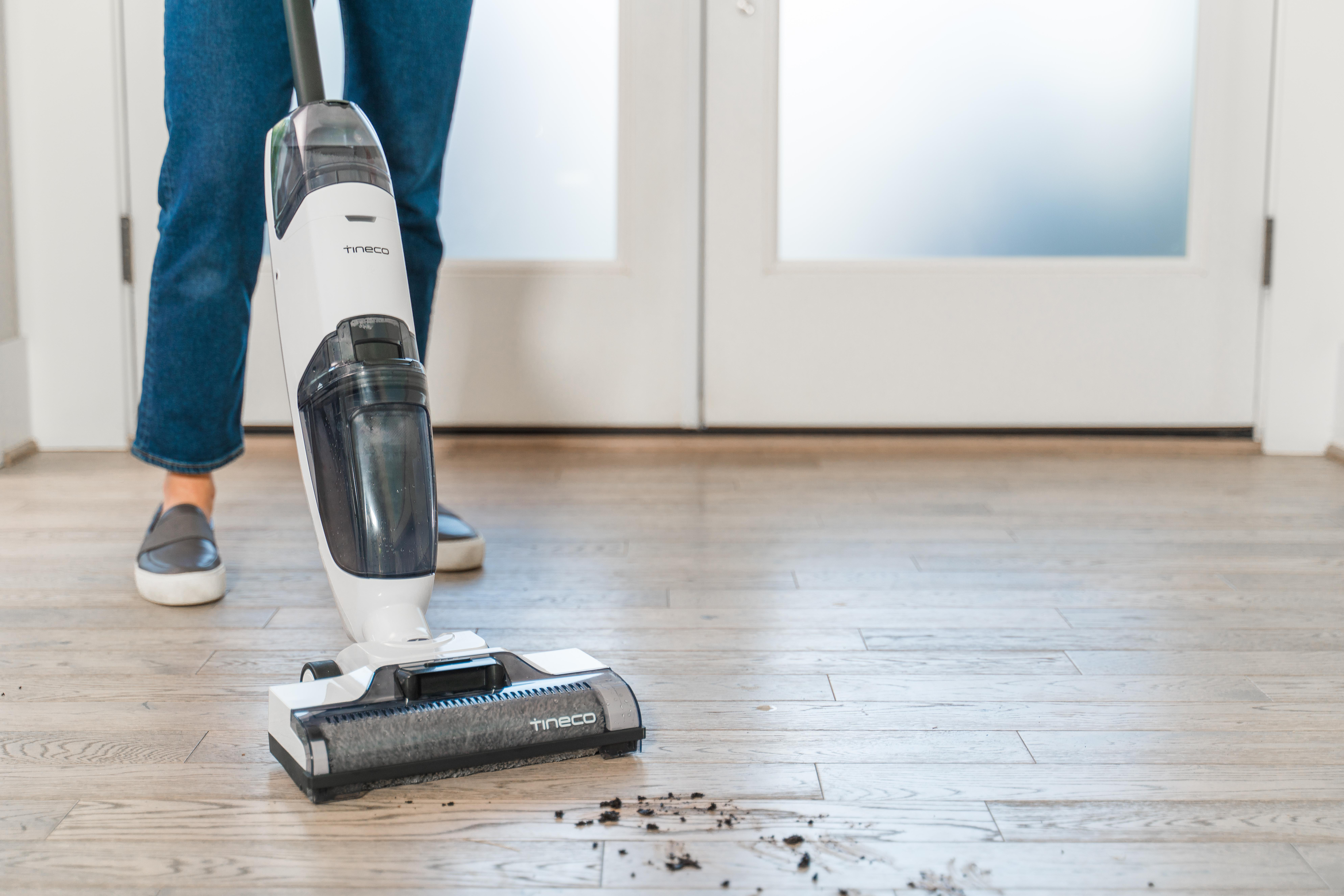 Eine Person benutzt einen weiß-schwarzen Staubsauger auf einem Holzboden mit Schmutz.