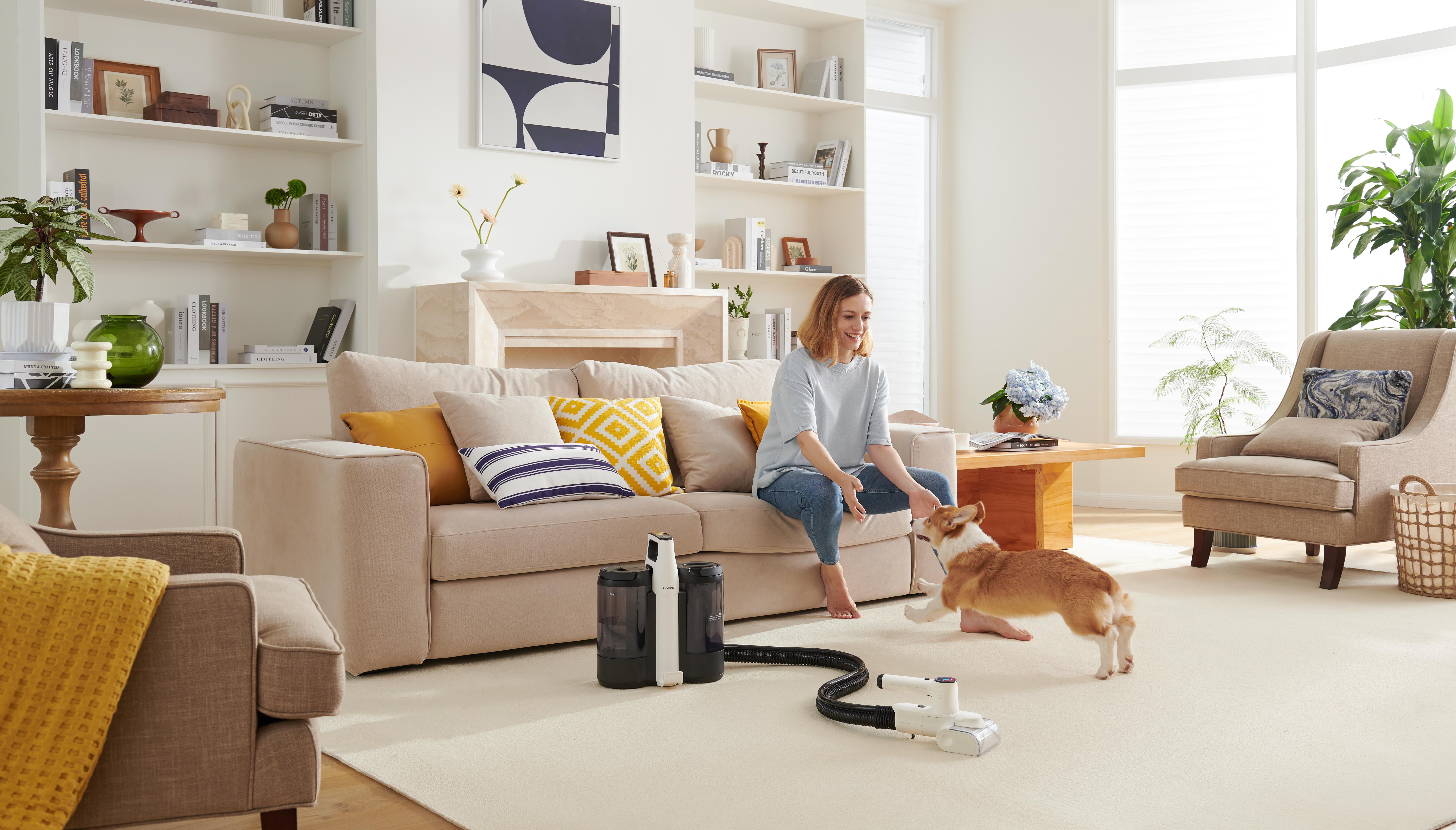 Eine Frau sitzt auf einem beigen Sofa und spielt mit einem Corgi. Ein Staubsauger steht in der Nähe auf dem Boden.