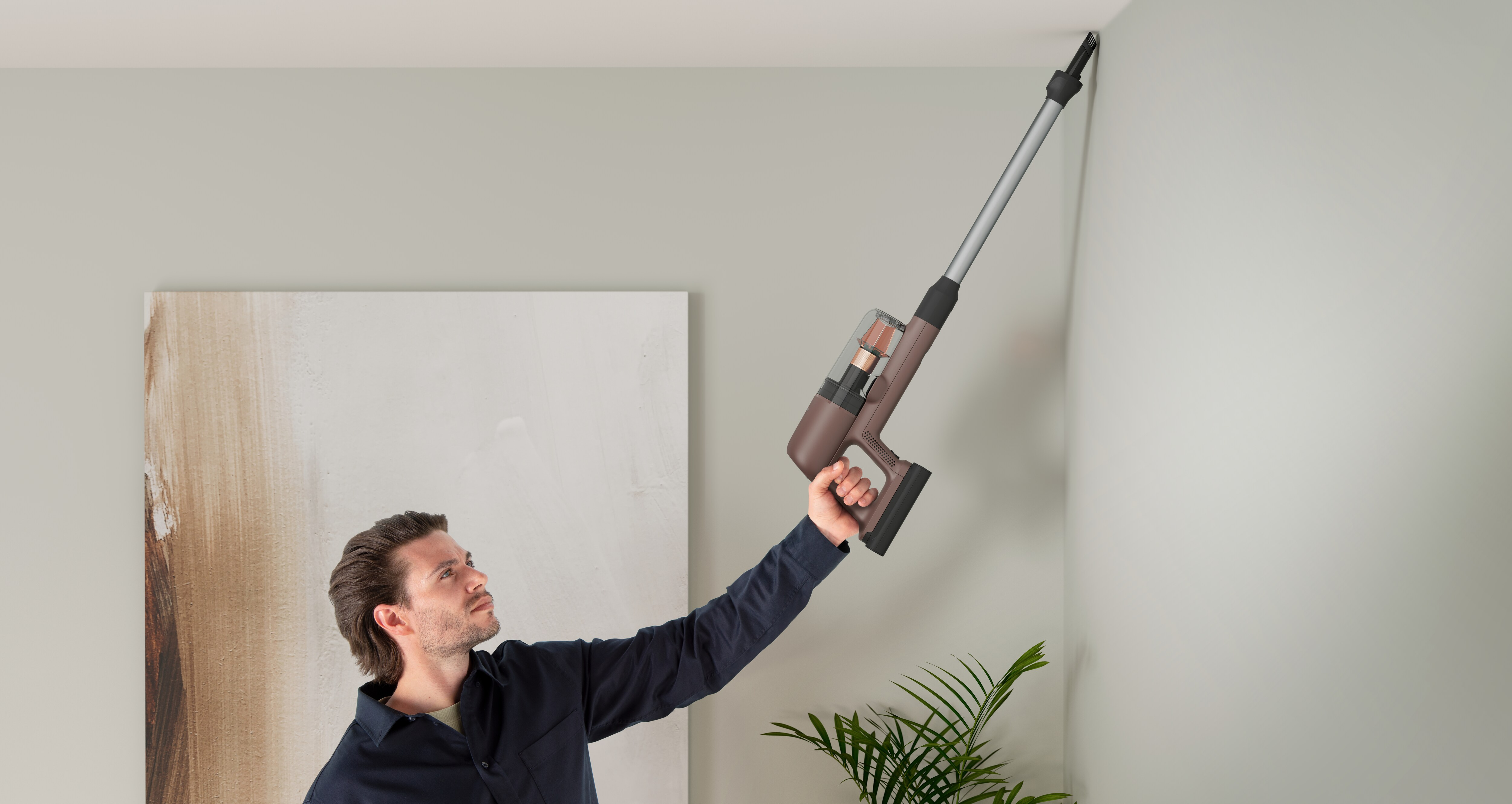 Ein Mann saugt mit einem Handstaubsauger die Decke ab. Der Raum hat neutrale Wände und ein Gemälde.