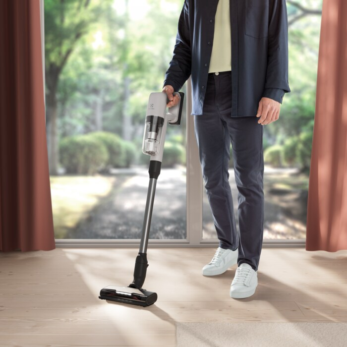 Un uomo aspira un pavimento in legno con un aspirapolvere a bastone bianco e grigio. Finestra e tende sullo sfondo.