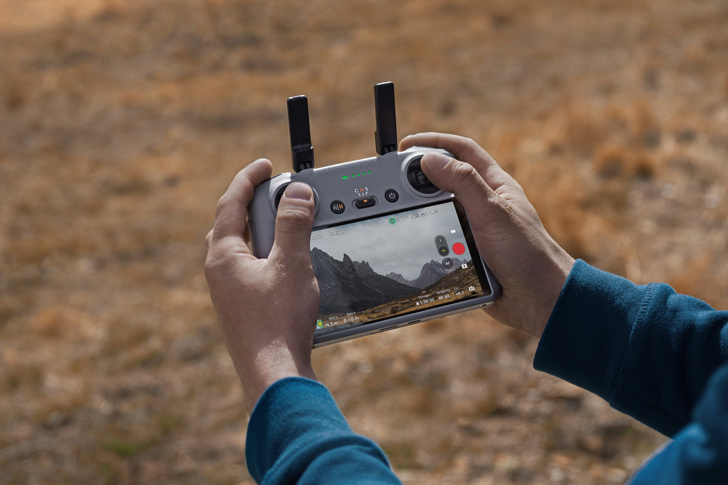 Hände halten Drohnen-Controller; Bildschirm zeigt Berglandschaft. Hintergrund verschwommenes Feld.