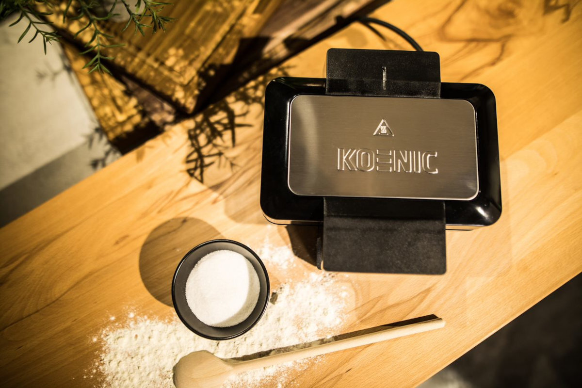 Widok z góry na gofrownicę, cukier i drewnianą łyżkę na drewnianej powierzchni. Mąka jest rozrzucona.