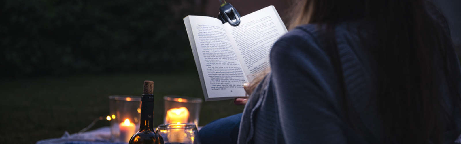 Person liest Buch im Freien, Kerzen und Weinflasche im Vordergrund, dunkler Hintergrund.