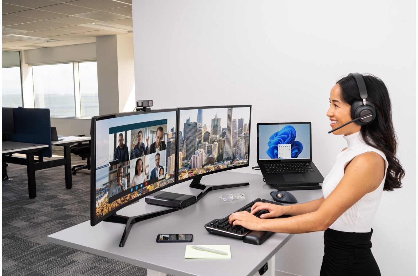 Egy nő dolgozik egy asztalnál két ívelt monitorral, laptopmal és fejhallgatóval. A helyszín egy irodai tér.