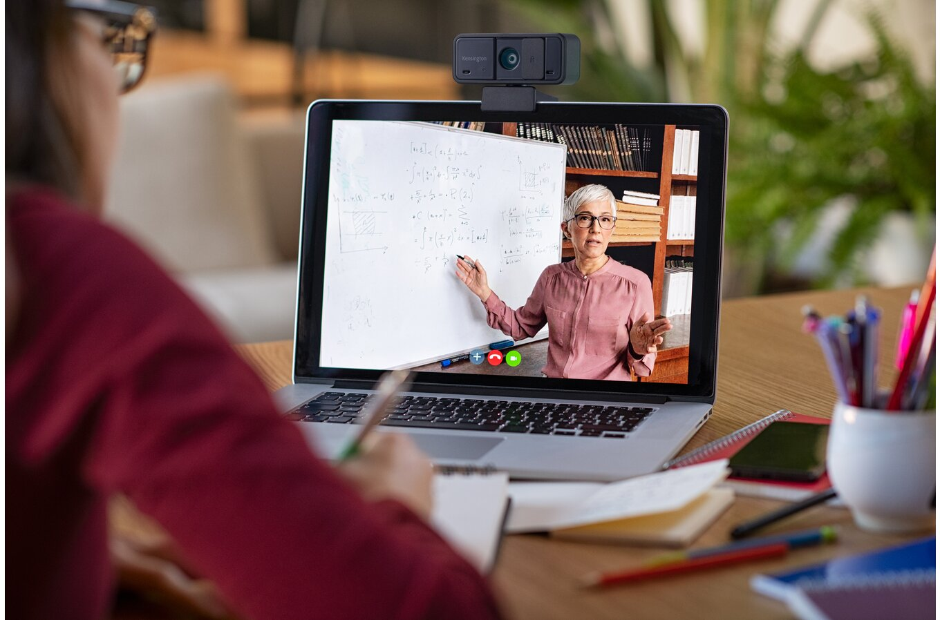 Nő laptopon videóhívásban egy tanárral. A képernyő egy táblát és egy osztálytermet mutat.