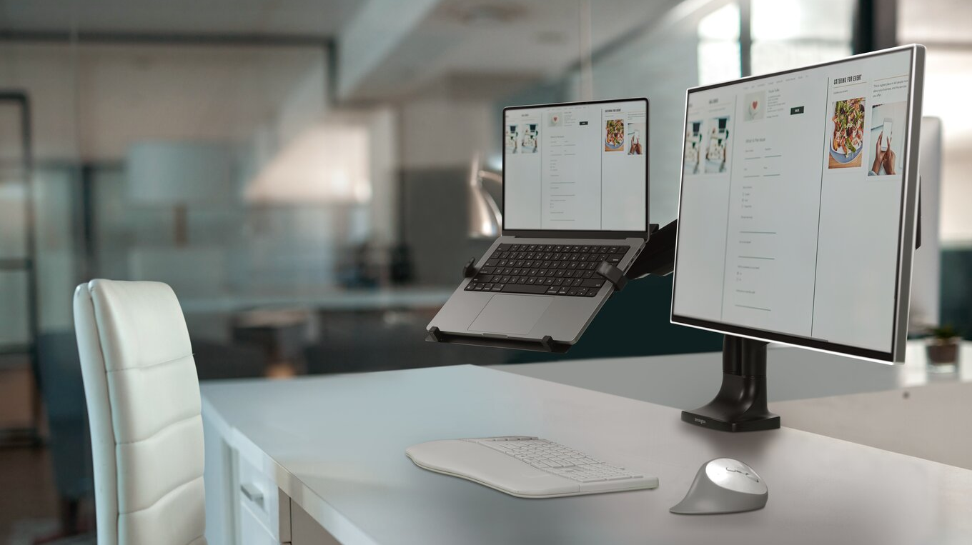 Egy munkaterület egy laptop állvánnyal, monitorral, billentyűzettel és egérrel. A háttér egy iroda.